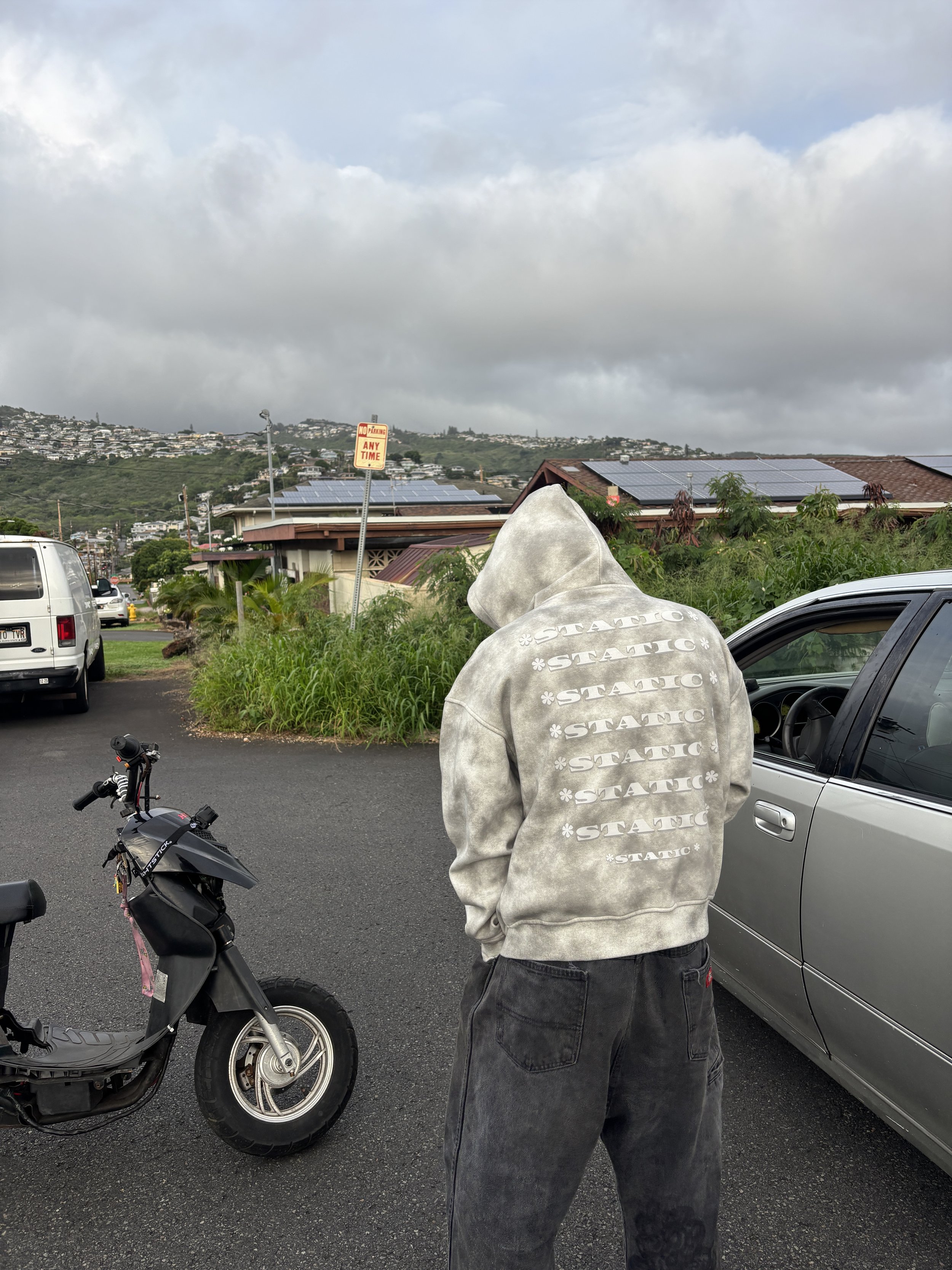Person wearing a gray hoodie with 'STATIC' written repeatedly on the back, standing in a parking lot near cars and a motorcycle, with a hillside and cloudy sky in the background.