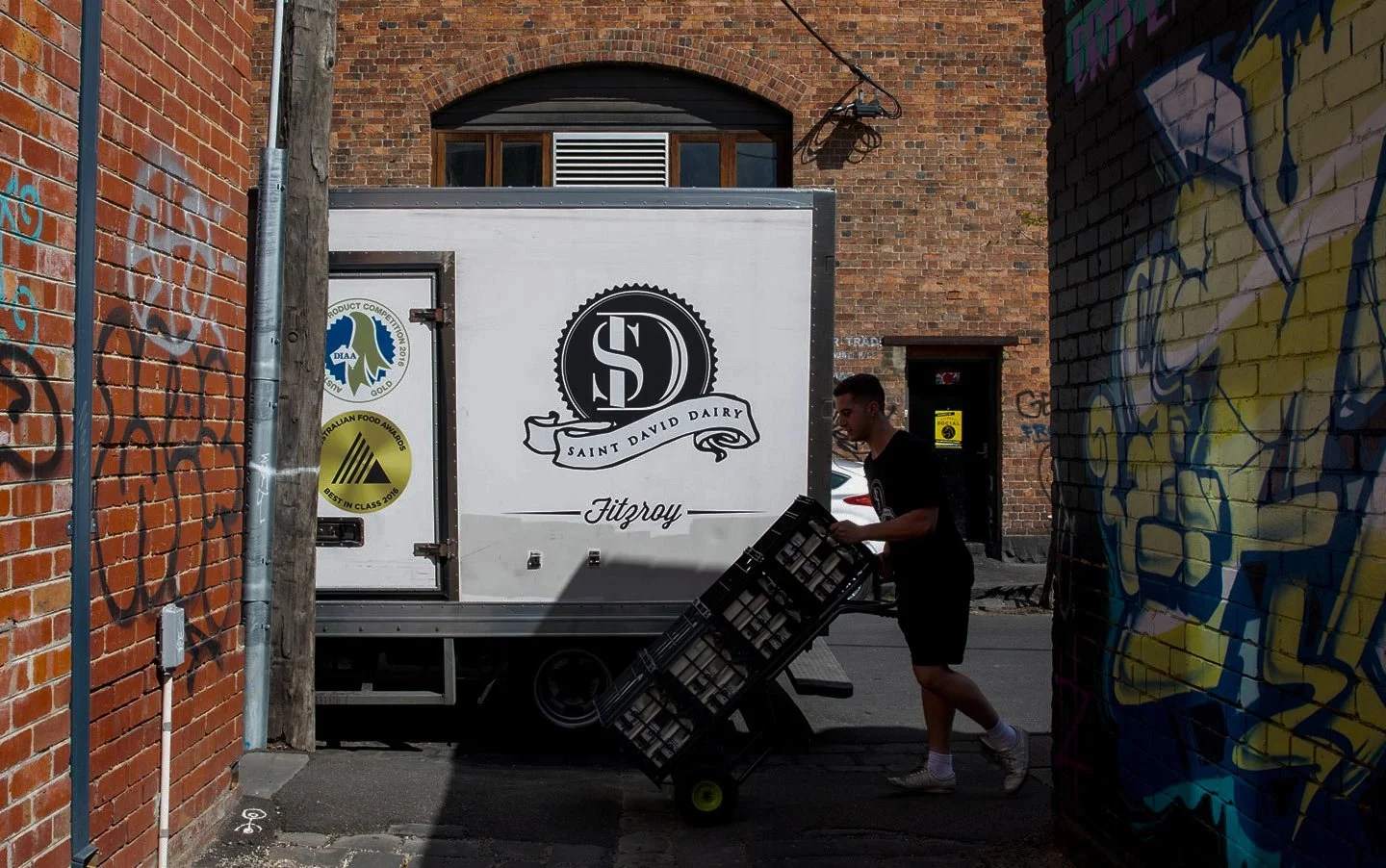 A person loading crates onto a truck with Saint David Dairy branding in an alleyway with graffiti-covered walls.