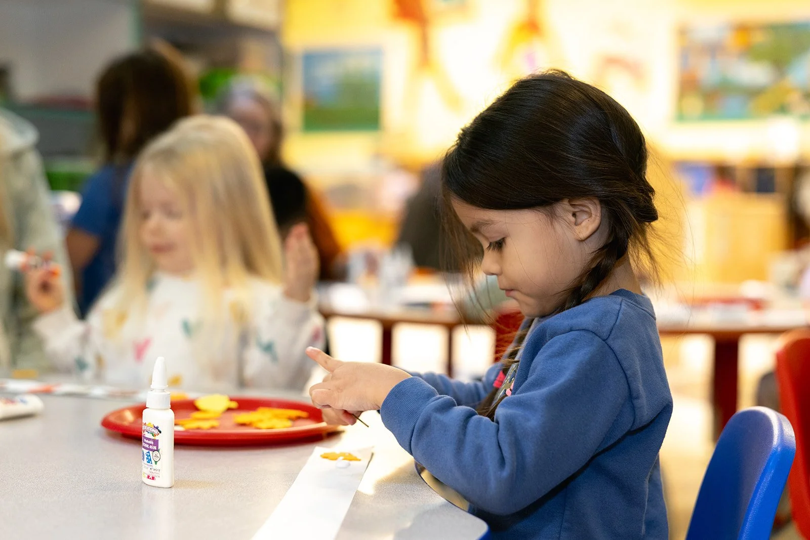 Child with dark haired pigtails wearing a blue shirt sitting at a table glueing down colored paper