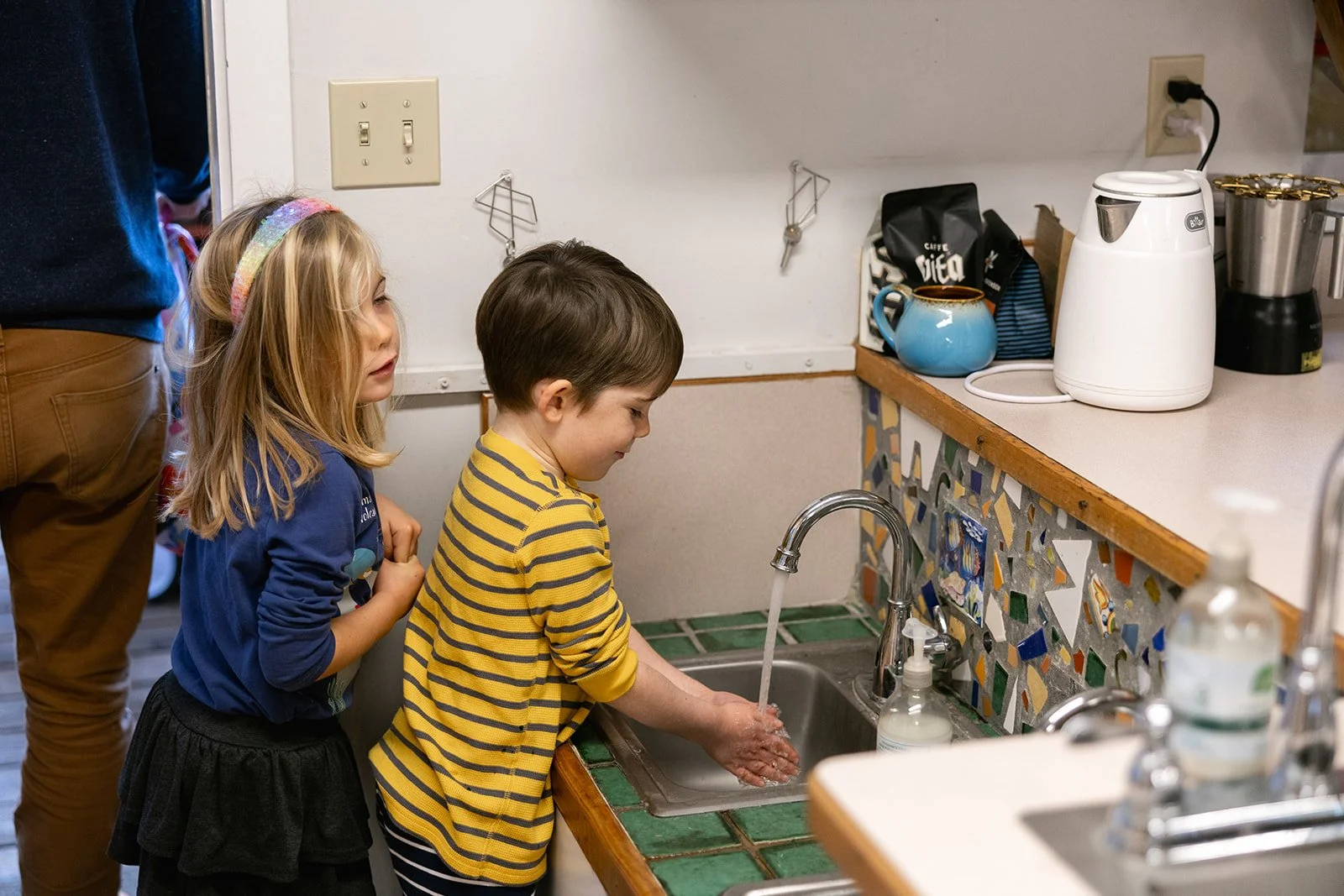 Child with short brown hair and yellow and blue striped shirt washing hand with another child with long blonde hair waiting behind them
