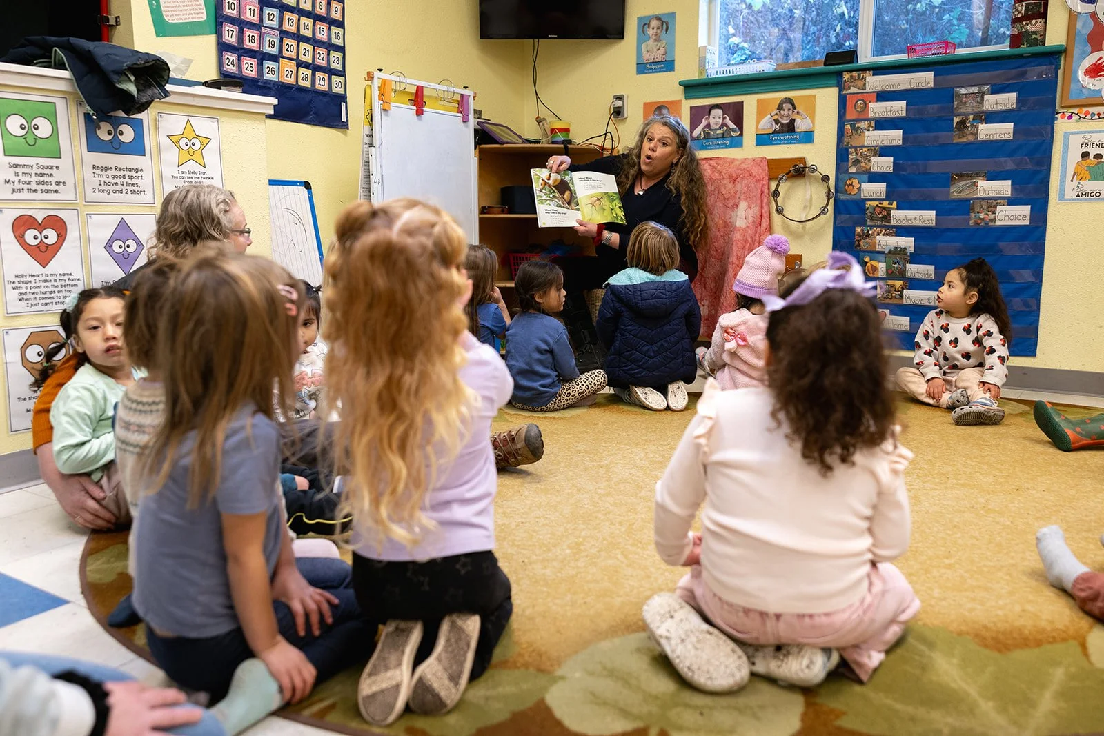 Teacher in front of classful of children sitting on the floor while they read for them