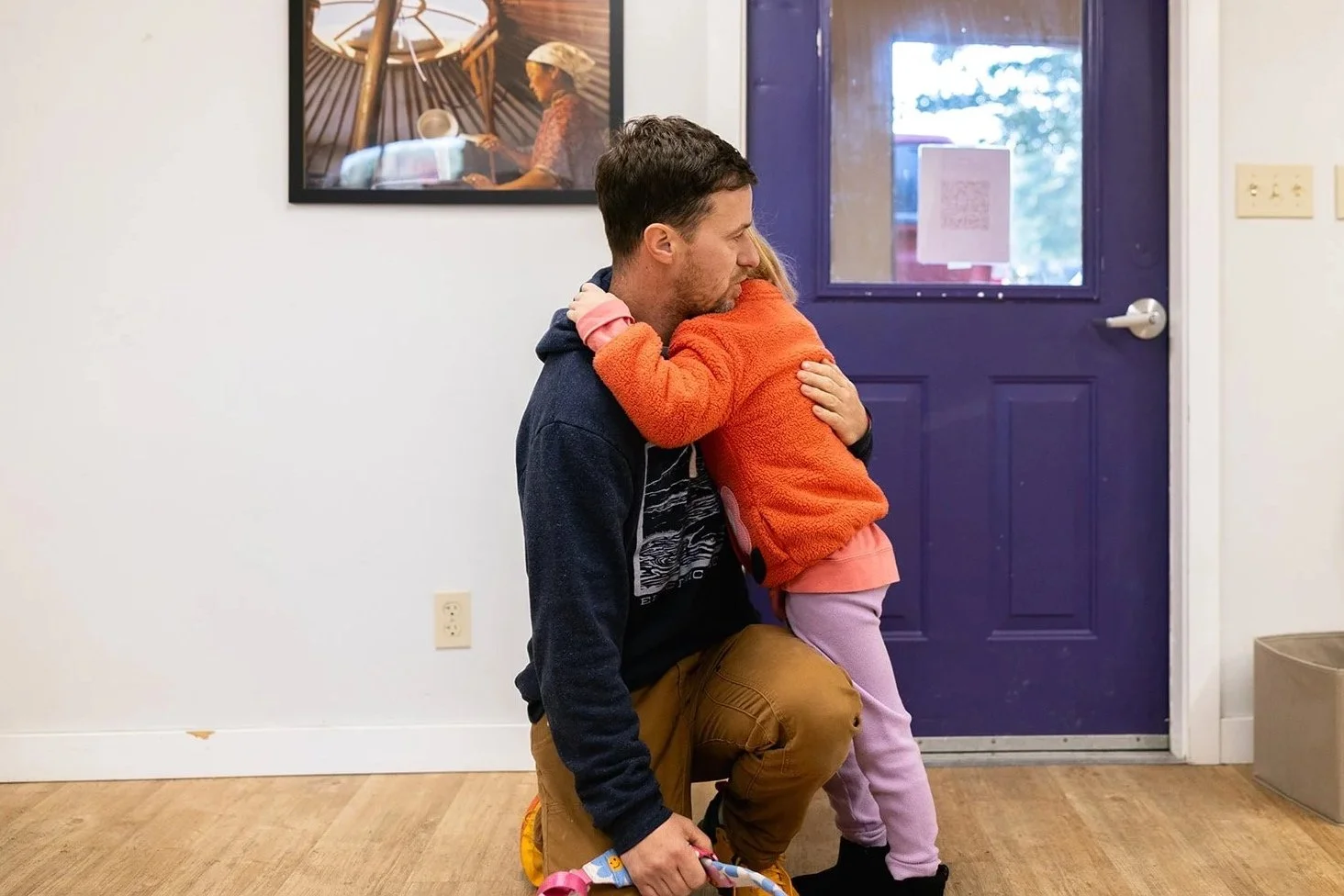 Parent kneeling hugging a child in an orange jacket with a purple door in the background