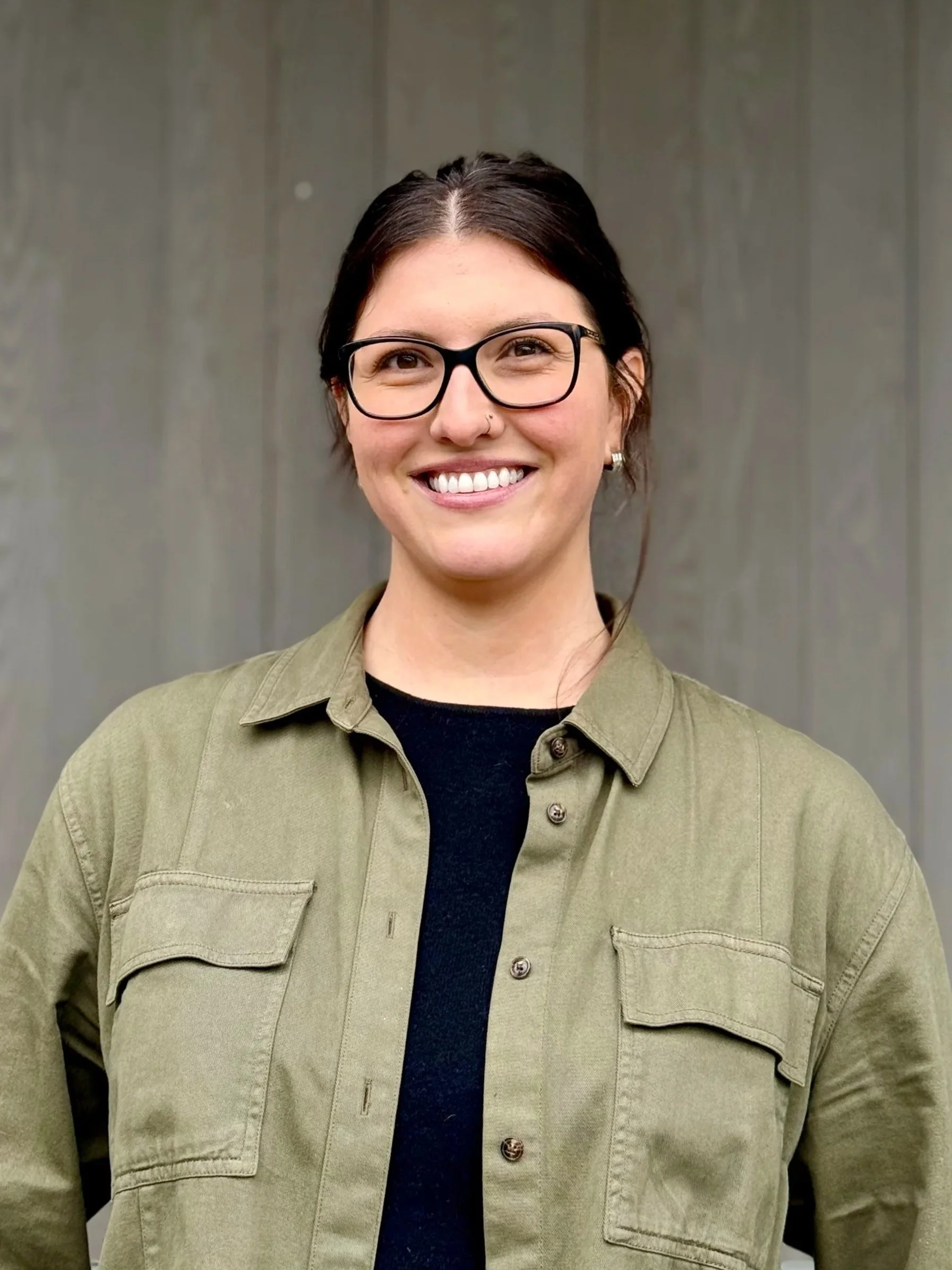 Portrait of person with dark hair pulled back and glasses wearing a dark shirt with green jacket