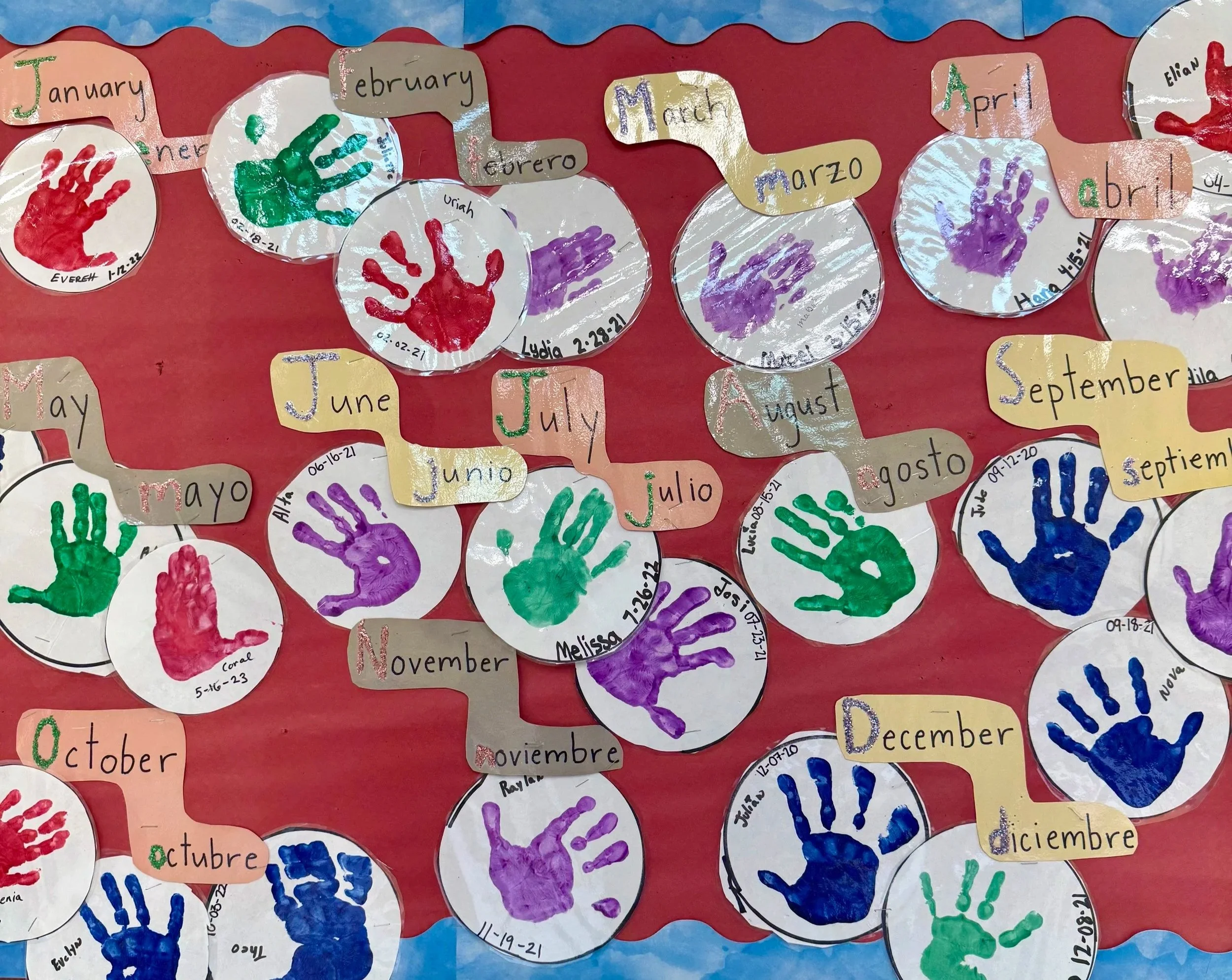 Different colored painted hand prints on while paper on a red background with the months of the year on a thumb tack board