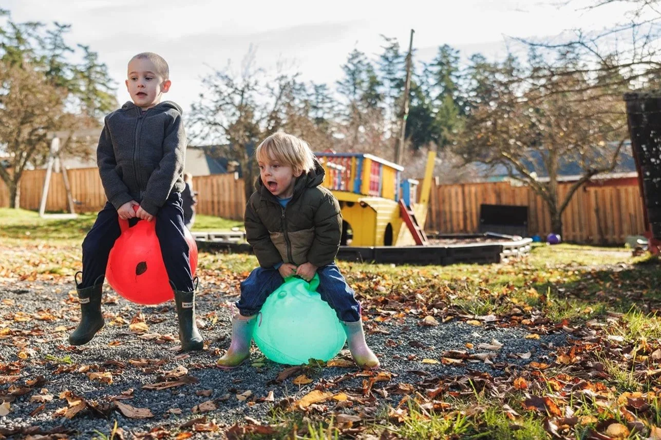 Two kids on bouncy balls. One is red. The other is green.  Wearing winter jackets and muck boots
