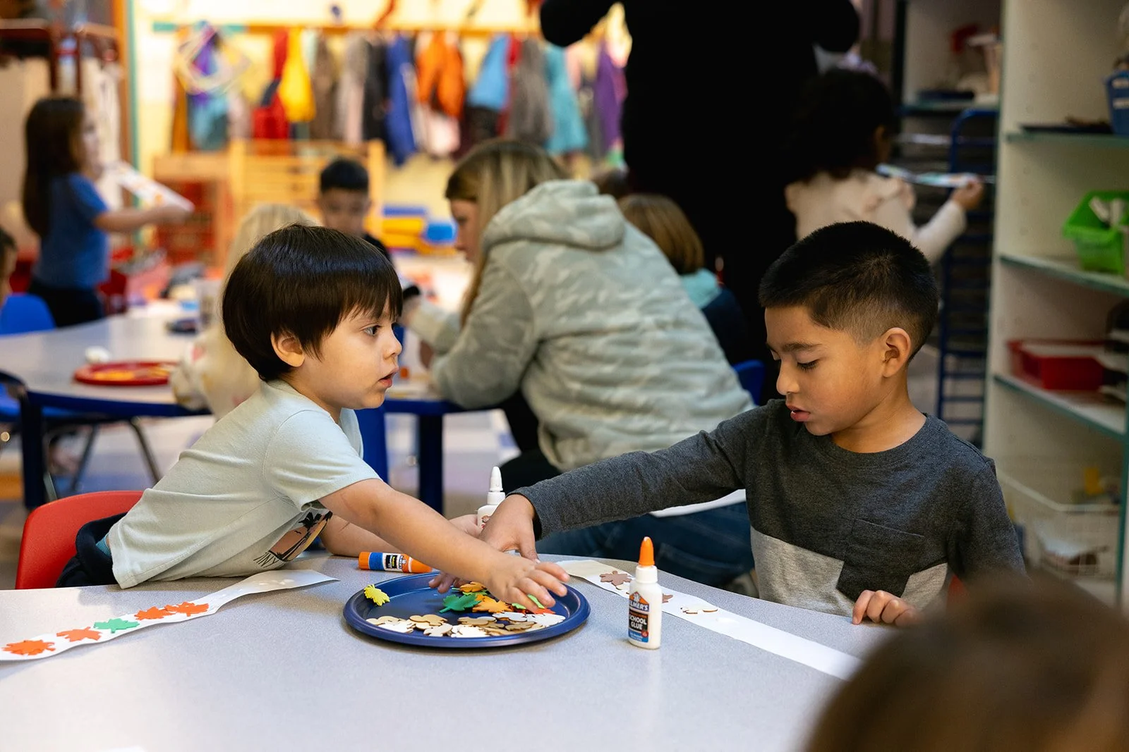 Two children sitting at a classroom table facing each other, gluing colored shapes to a strip of paper