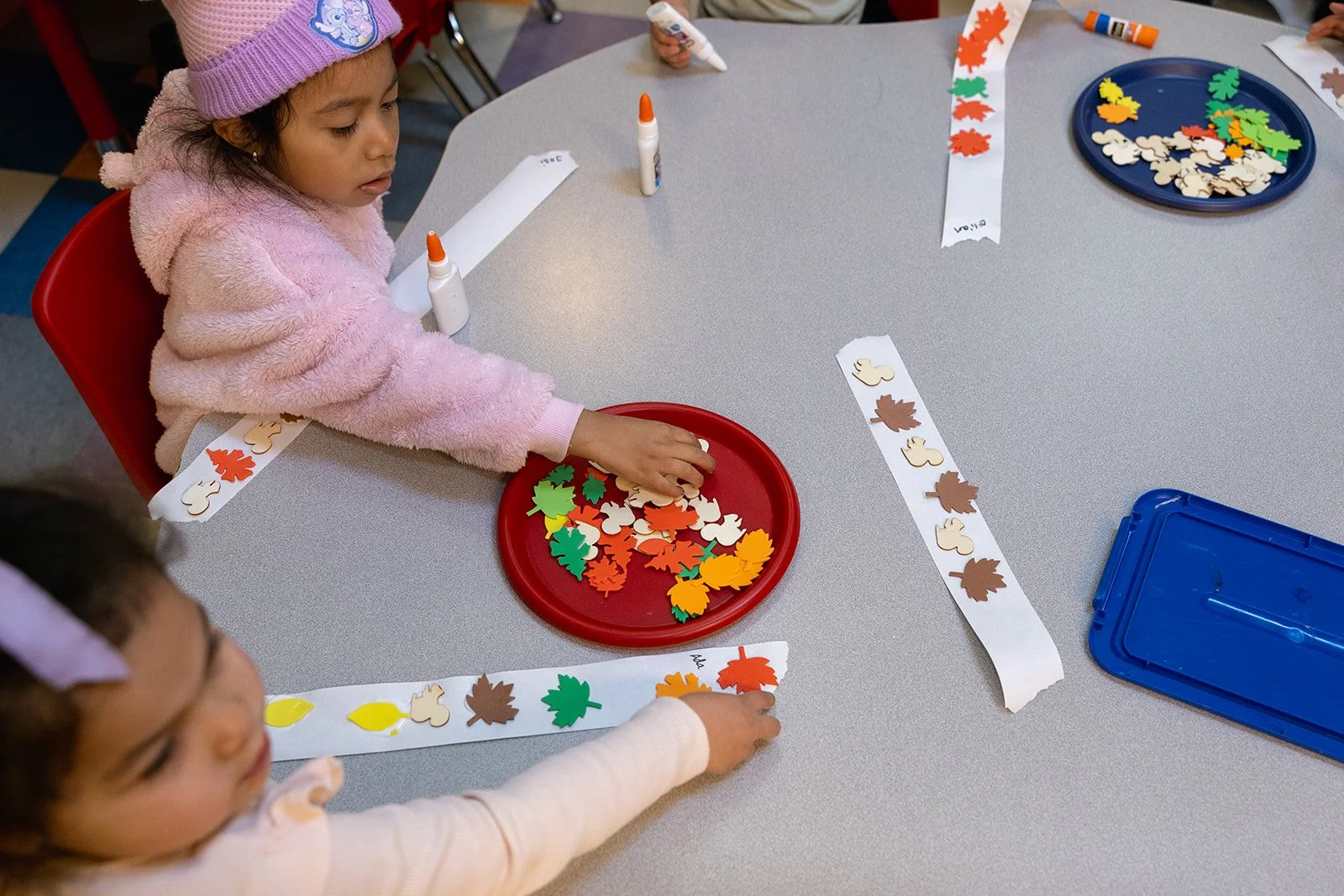 Two kids from above sitting a table with plate of different colored paper leaves