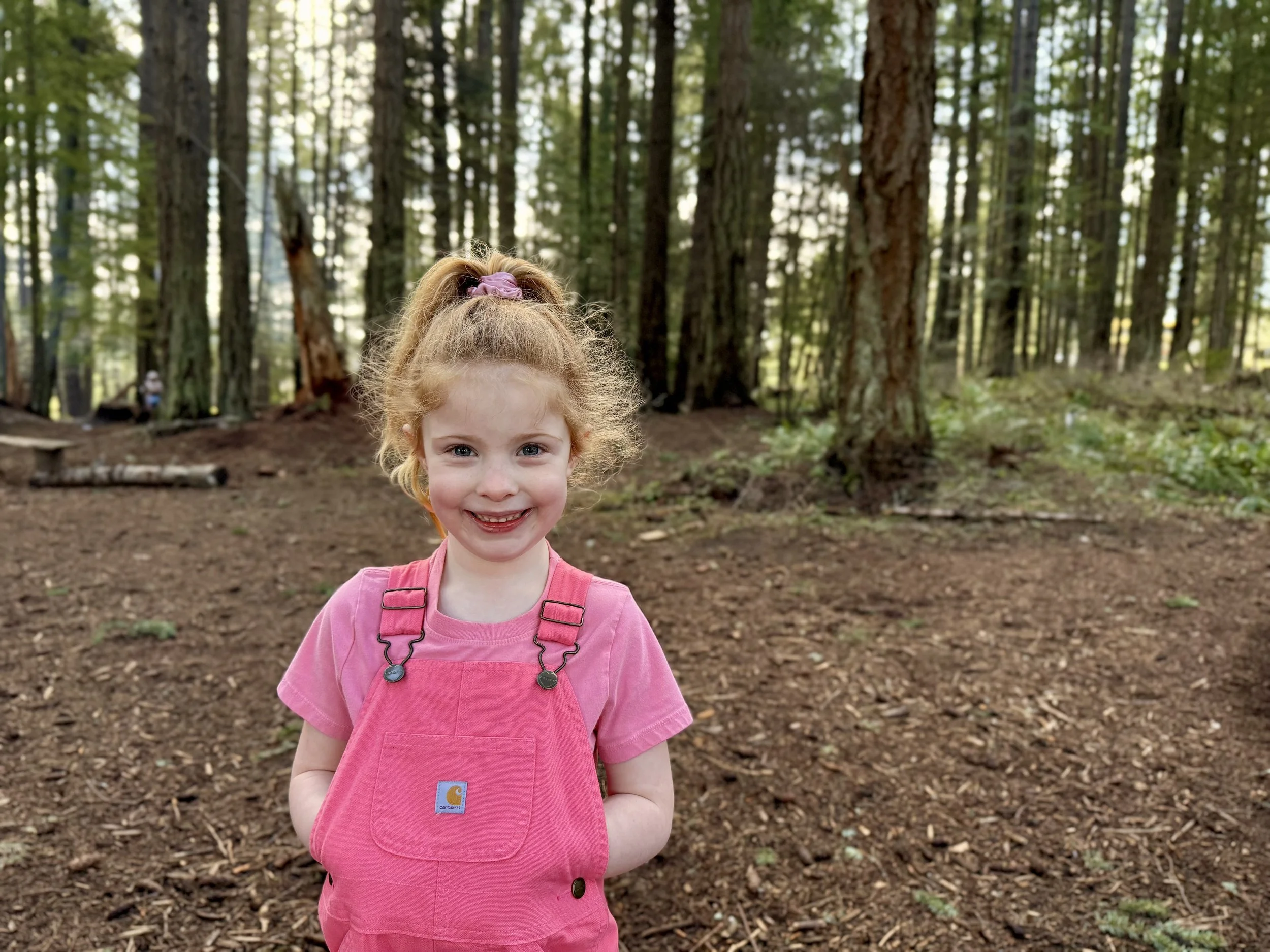 Child with hair in pony tail wearing a pink t-shirt and overalls smiling at camera while in the woods