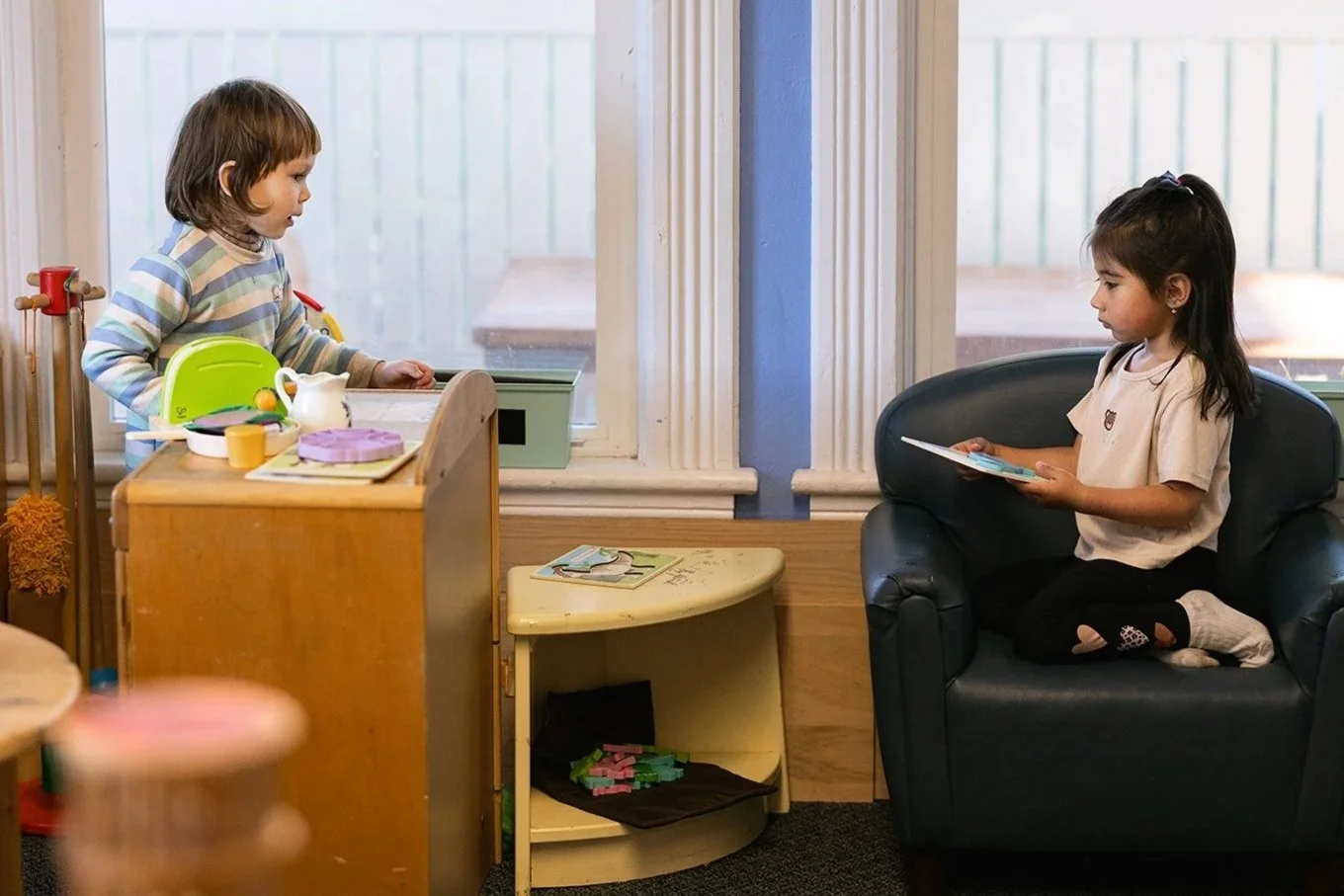 Two children from the side looking at each other. One siting in a chair reading a book. The other standing in front of a table with toys on it.