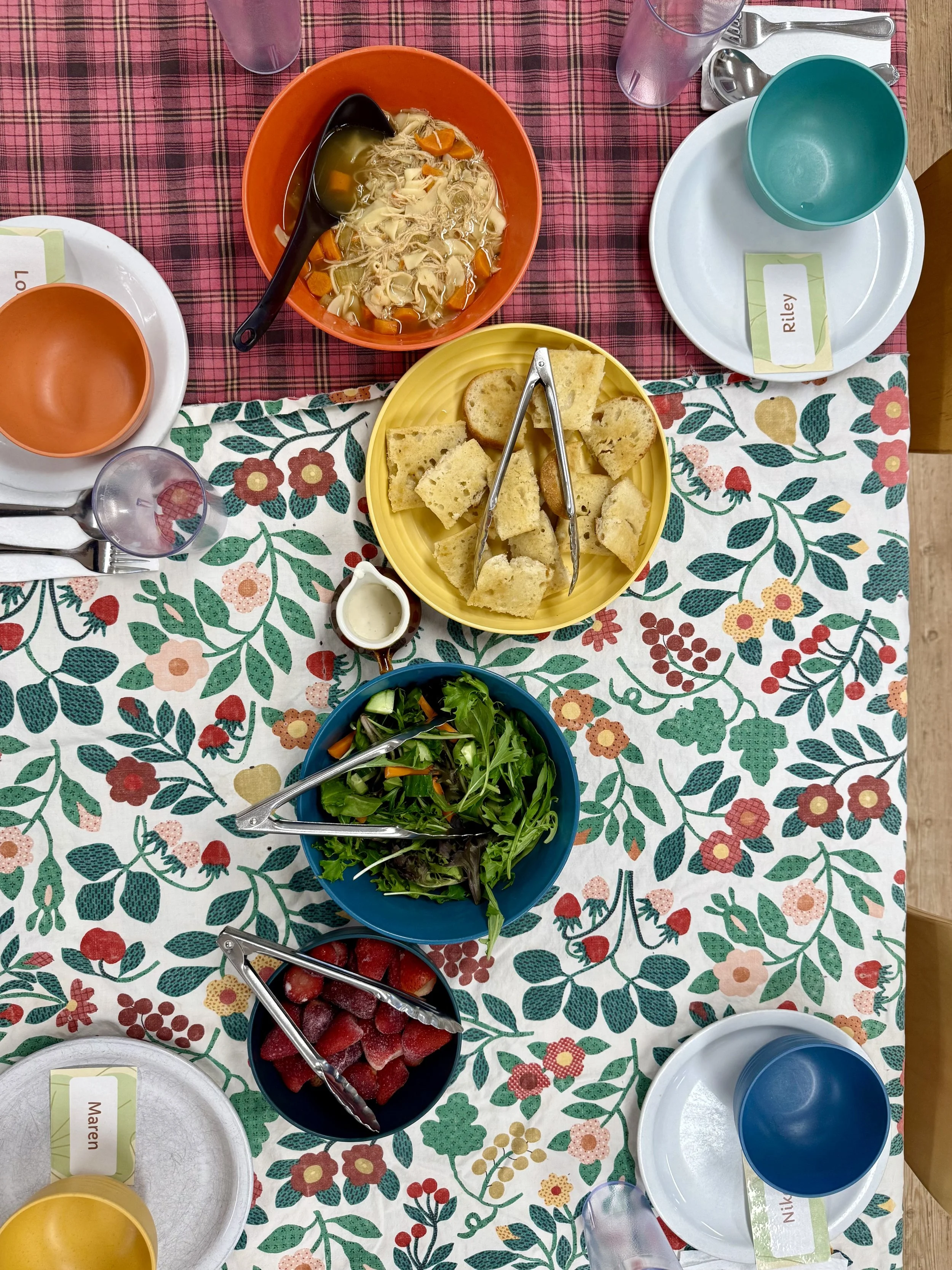 A table from above with two patterned table cloths and colored bowls of food with serving utensils.