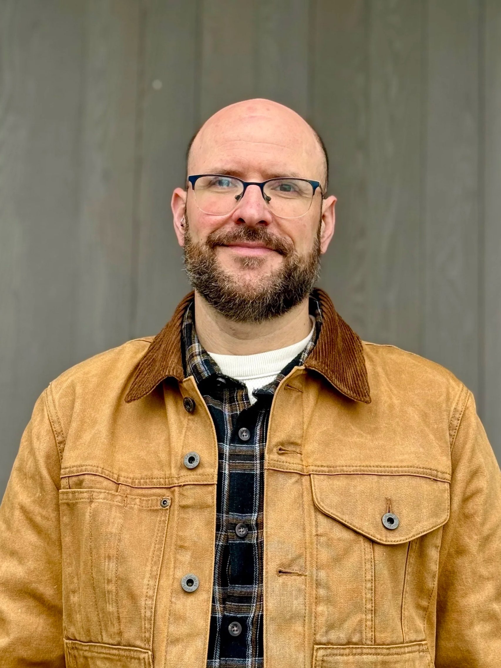 Portrait of person with no hair on head, beard, and mustache wearing glasses with a tan jacket and plaid shirt