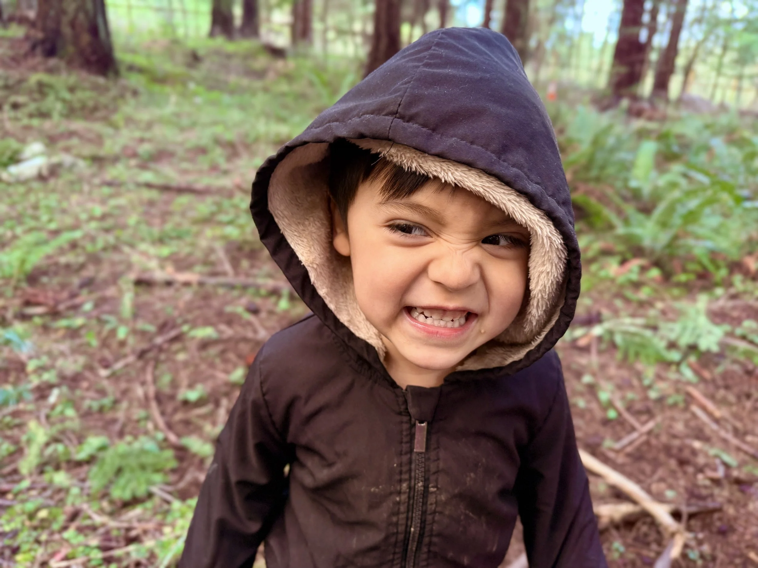 Kid outside wearing a dark hooded jacket making a funny face showing his teeth
