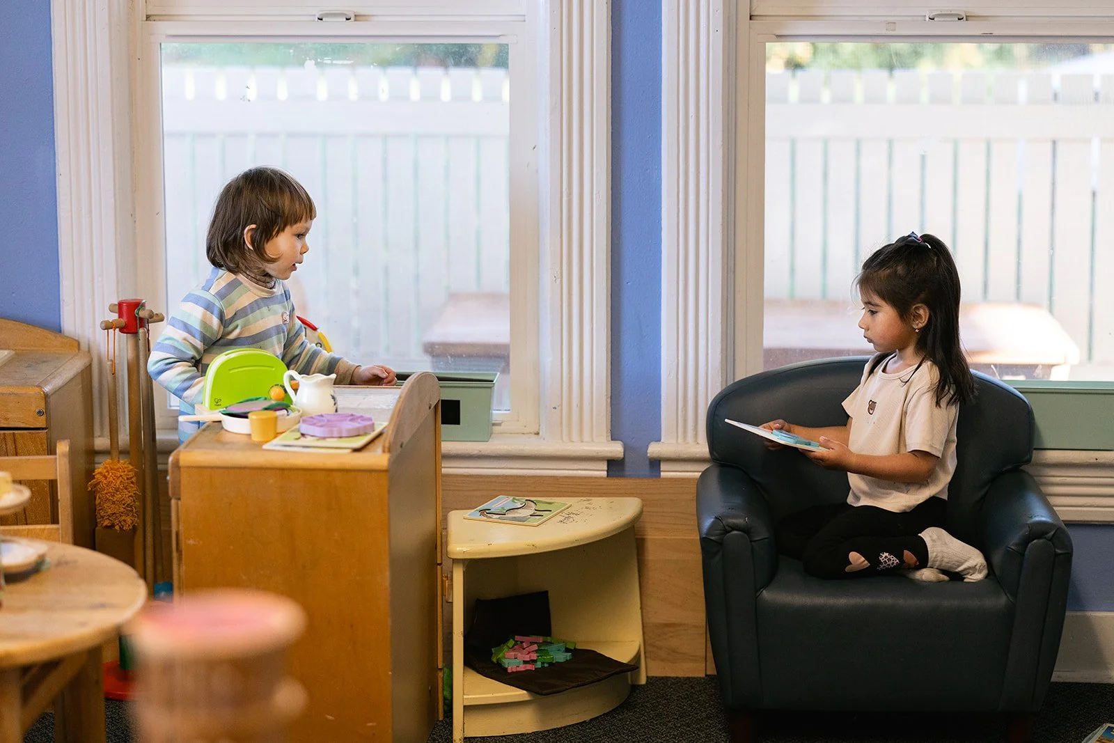 Two children from the side looking at each other. One siting in a chair reading a book. The other standing in front of a table with toys on it.
