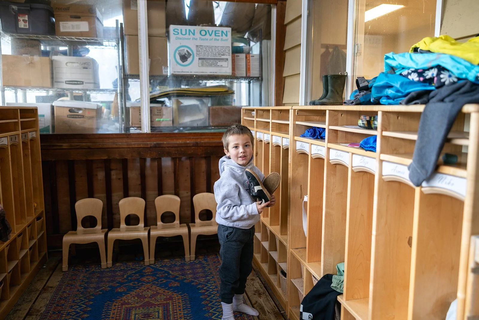 Child looking at camera with grey hoodie and shoes off standing in front of cubbies