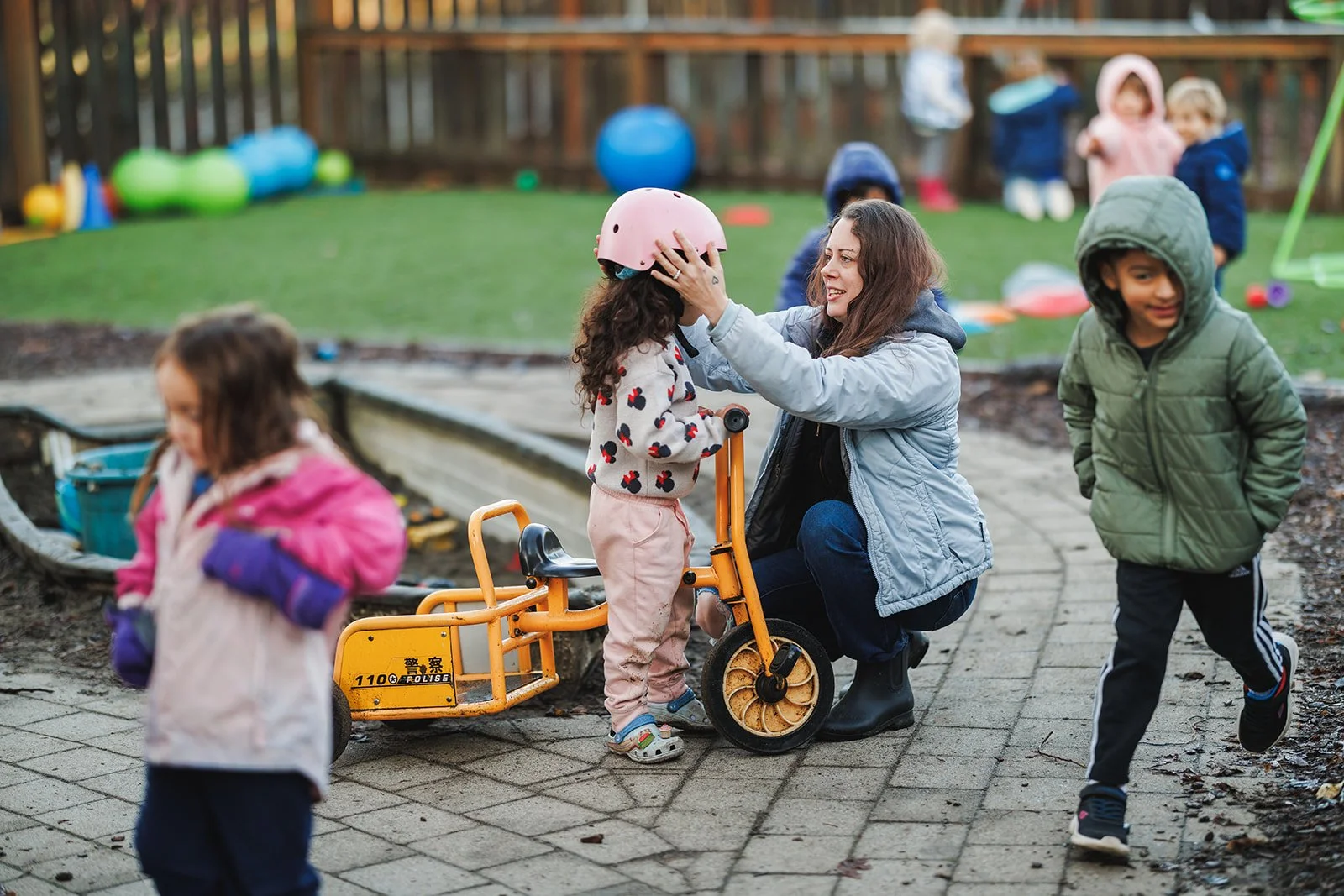 Teacher is light blue jacket putting pink bike helmet on child
