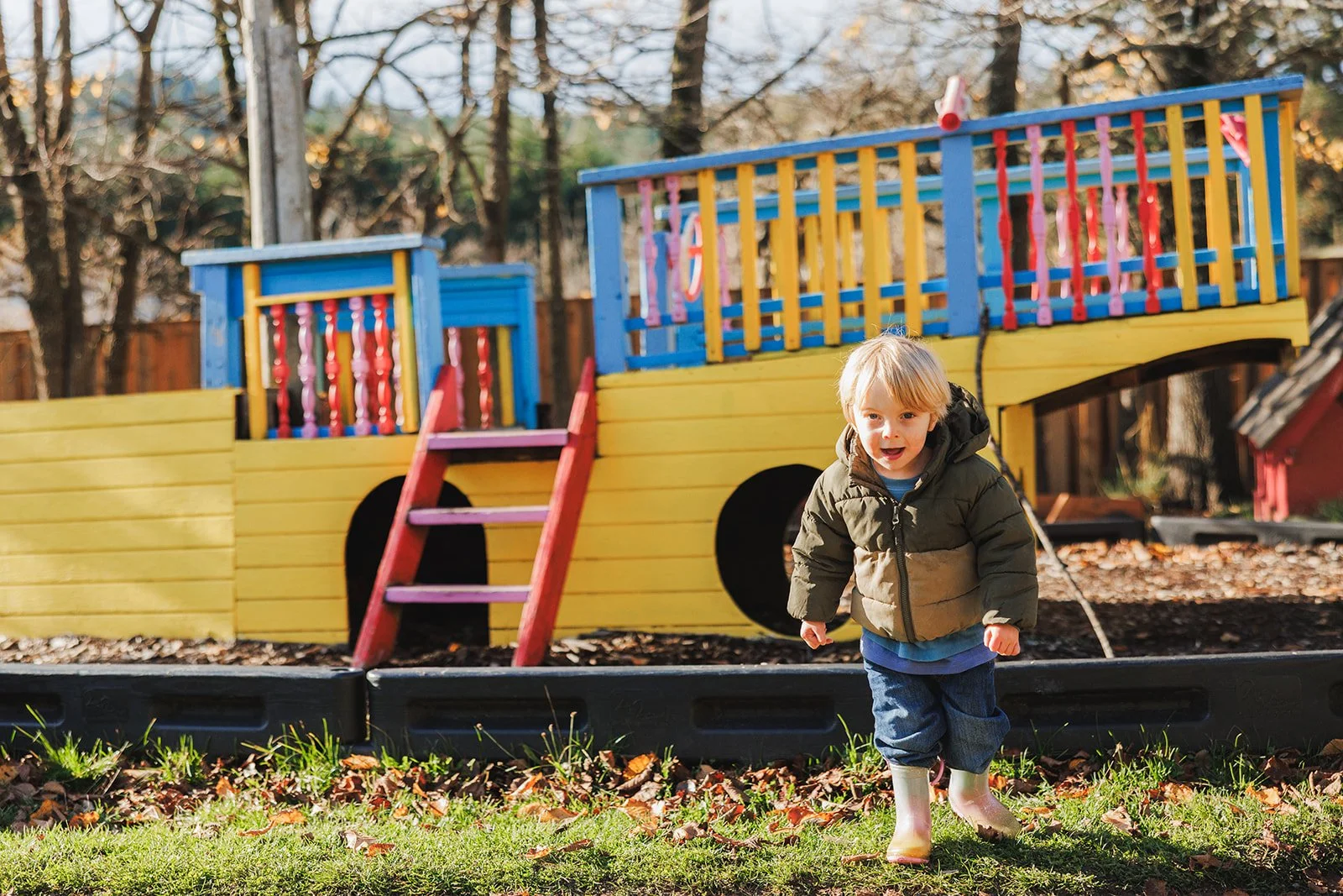Kid in with blond hair and brown jacket running towards camera with ship-like play structure in background