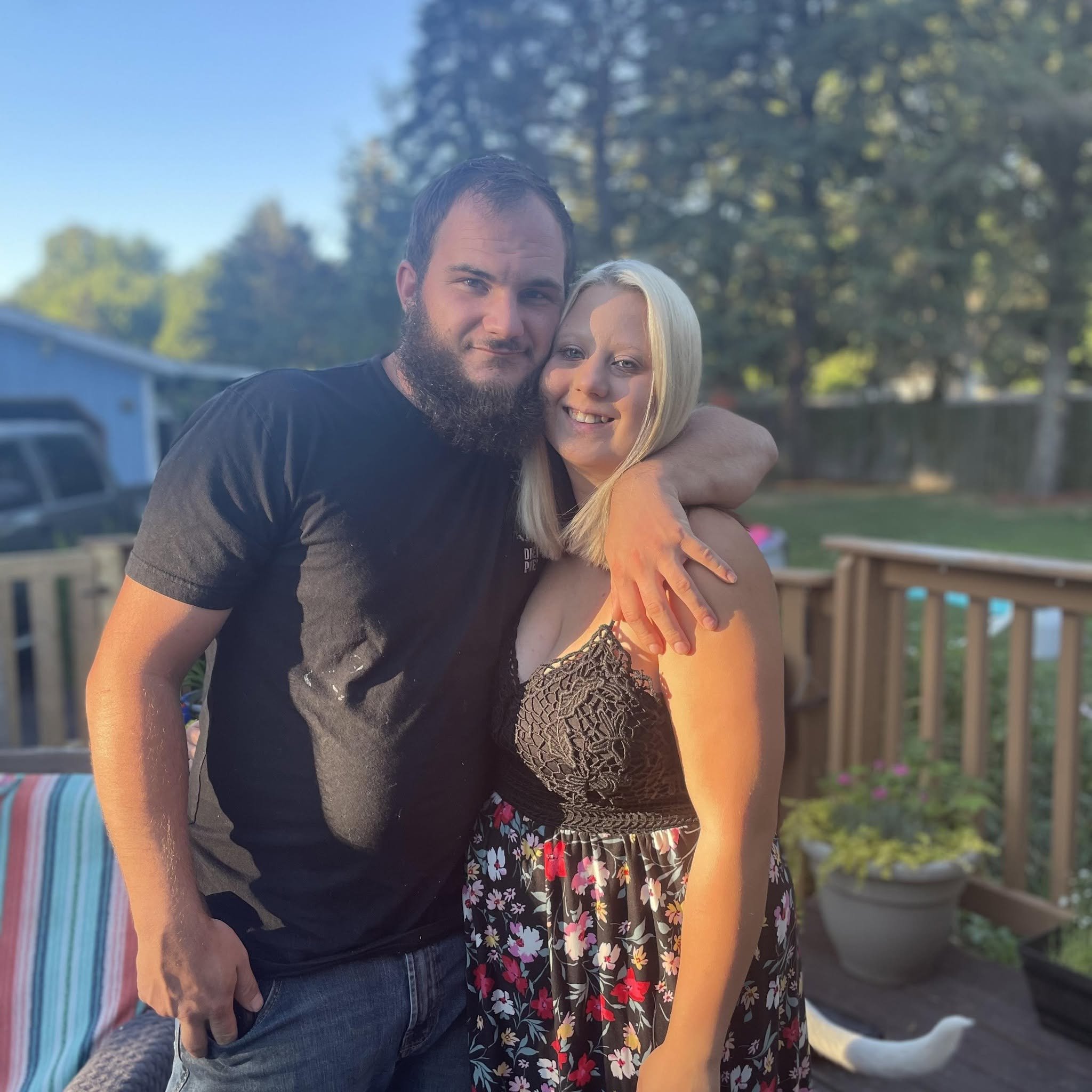 A man with a beard and a woman with blonde hair are hugging outdoors, smiling at the camera, with trees and a wooden fence in the background during sunset.