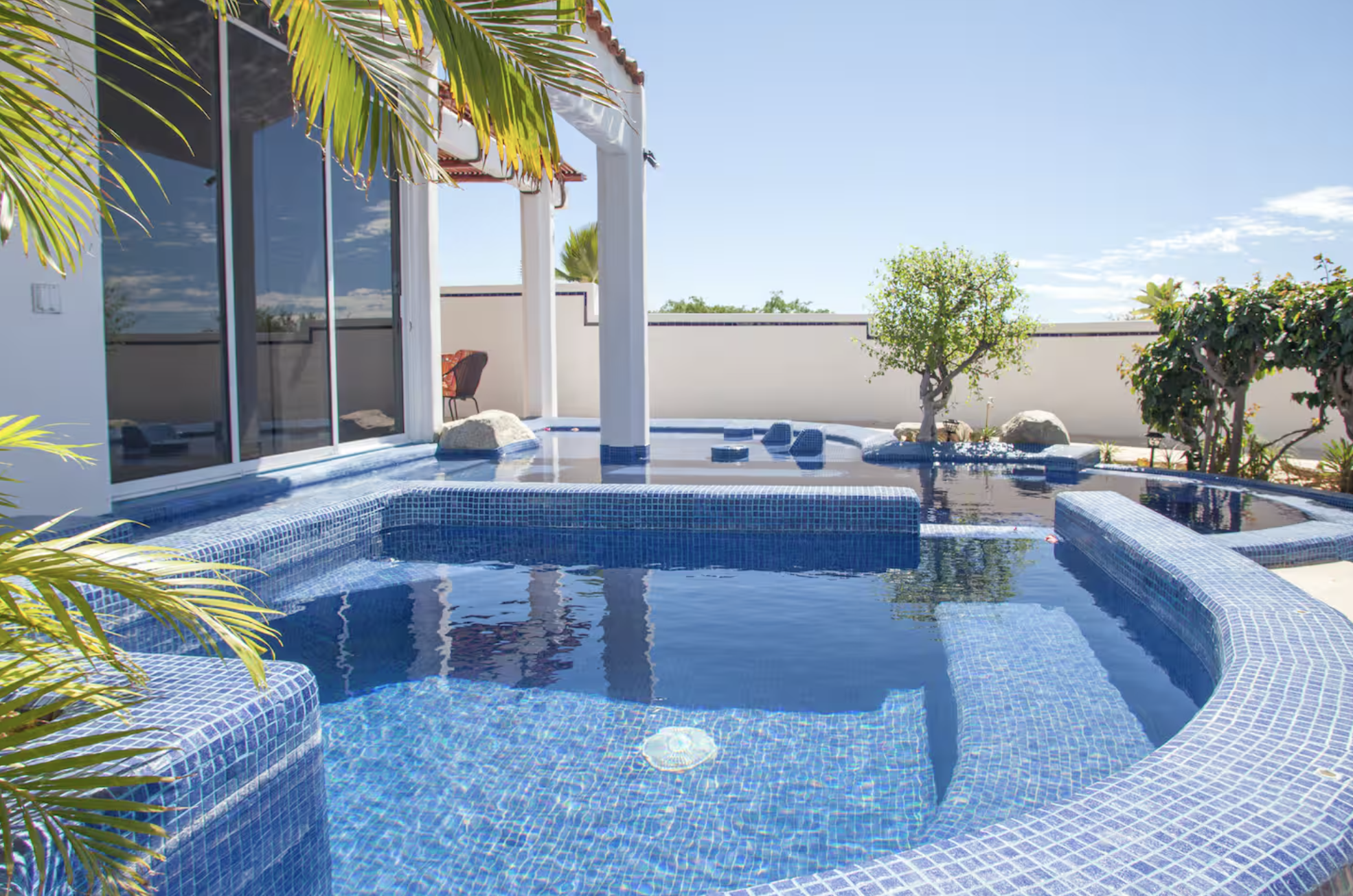Luxury outdoor pool with blue mosaic tiles, surrounded by lush greenery, in a sunny backyard with a private patio and glass doors leading inside.