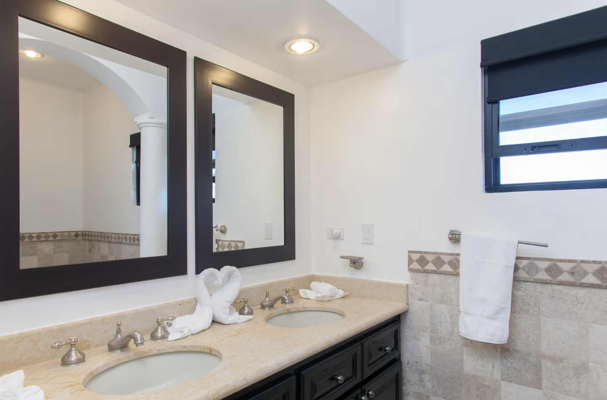 Bathroom with dual sinks and mirrors, towel rack with hanging towel, window with blinds, and towel folded into swan shapes.