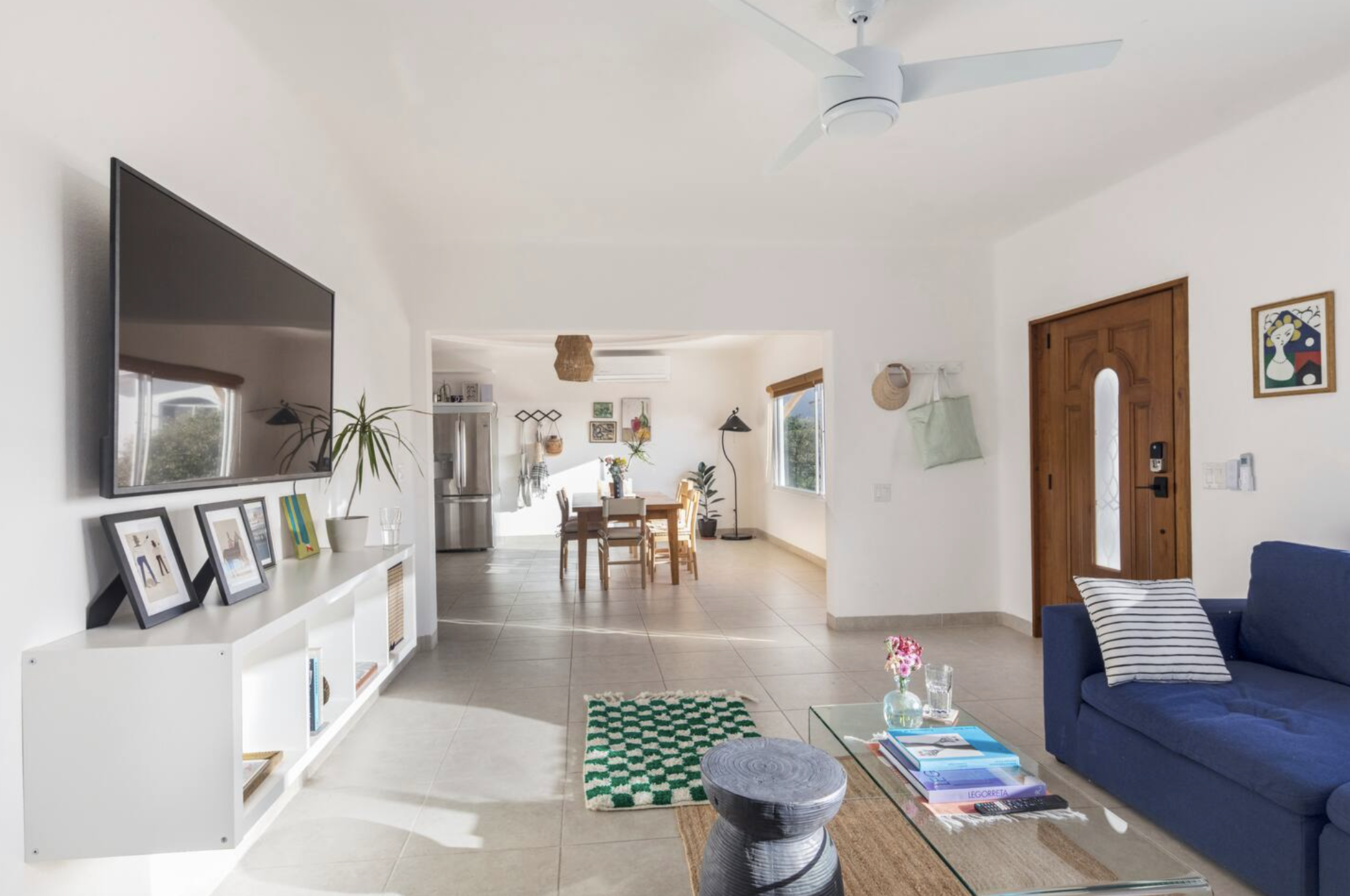 Living room with a blue sofa, glass coffee table with books and flowers, mounted TV, white console with framed photos, and a view into the dining area with a wooden table, chairs, and artwork on the walls.