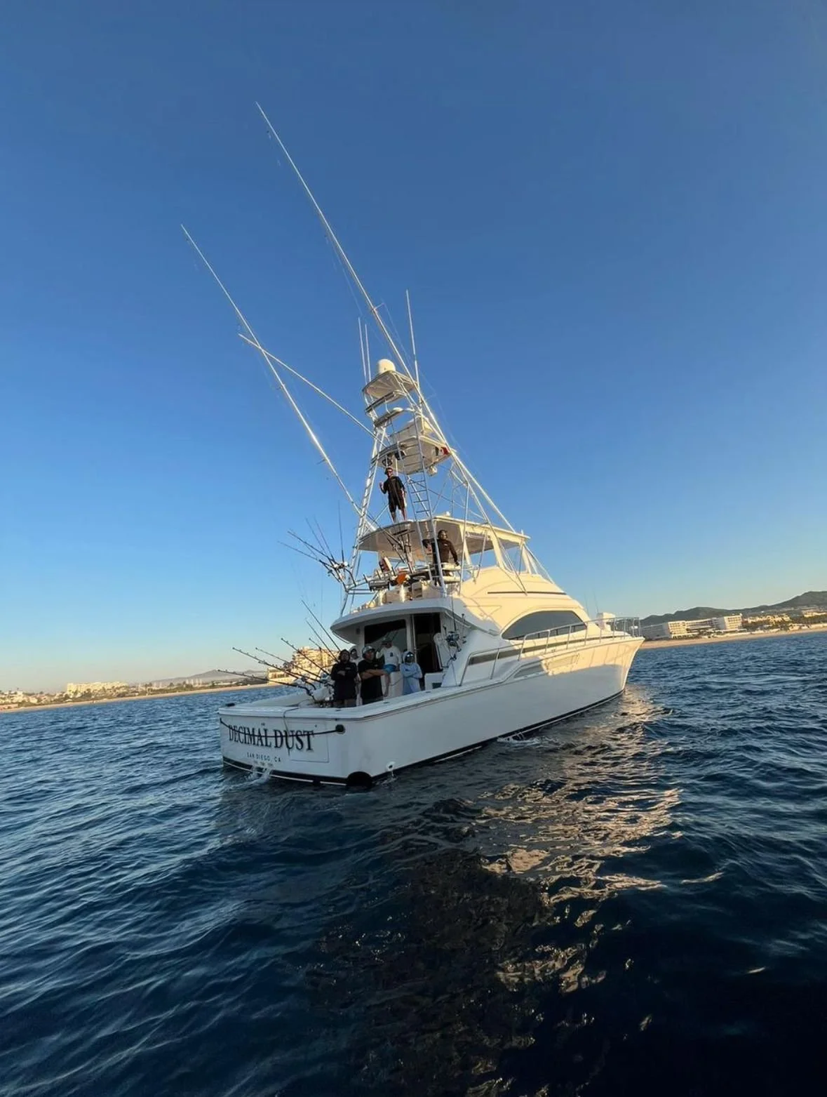 A white fishing boat named 'BEACH BAL DUST' on the water with people onboard, some fishing rods on the side, and a clear blue sky in the background.