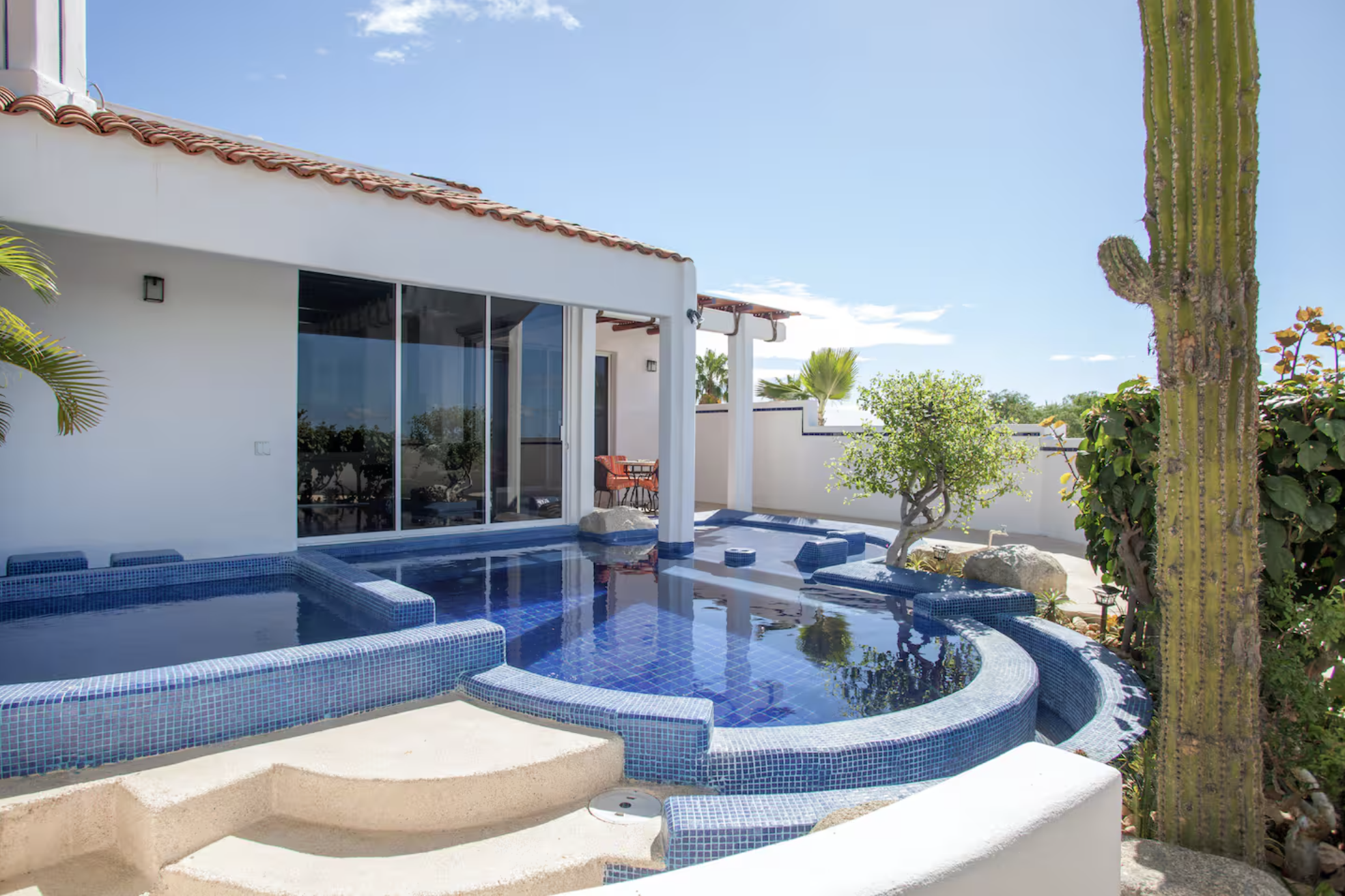 Modern backyard with a small blue-tiled pool, spa, potted plants, and a cactus, with a white house featuring sliding glass doors and patio furniture under a clear blue sky.