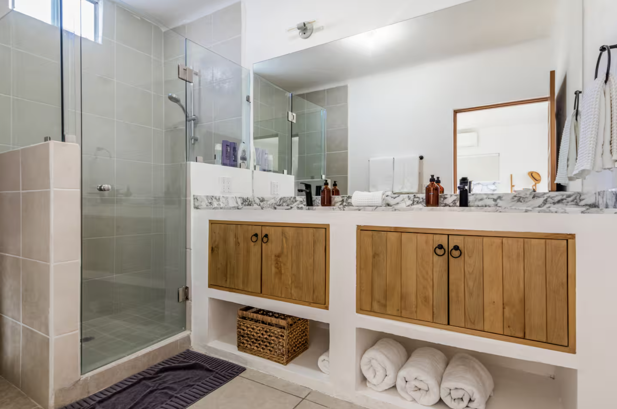 Bathroom with glass shower enclosure, double vanity with marble countertop, and wooden cabinet doors, with rolled towels and toiletries.