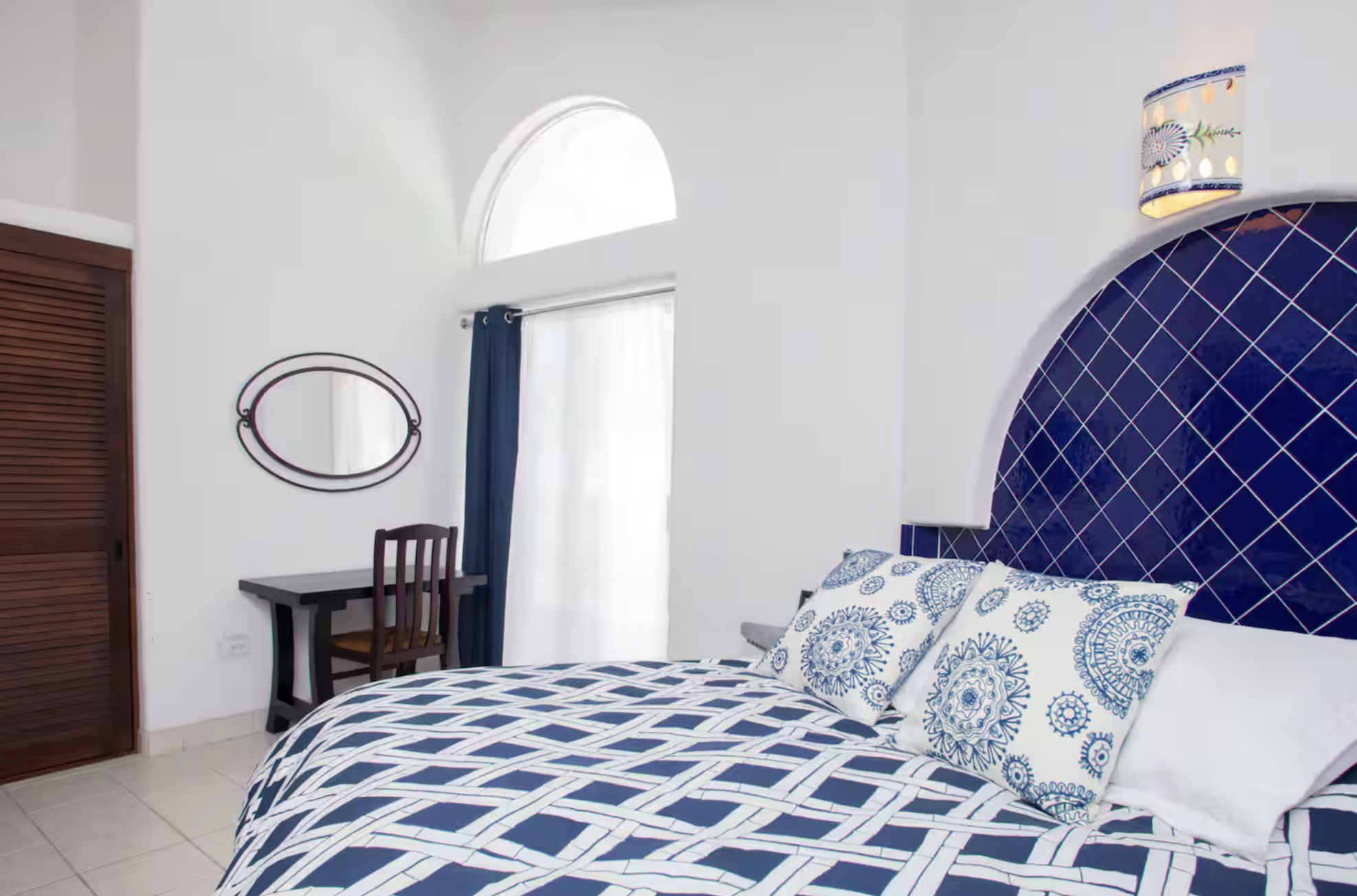Bedroom with a bed, blue and white bedding, white walls, arched window, blue curtains, dark wood desk, chair, and mirror, round wall light, wooden closet, and a decorative lamp.