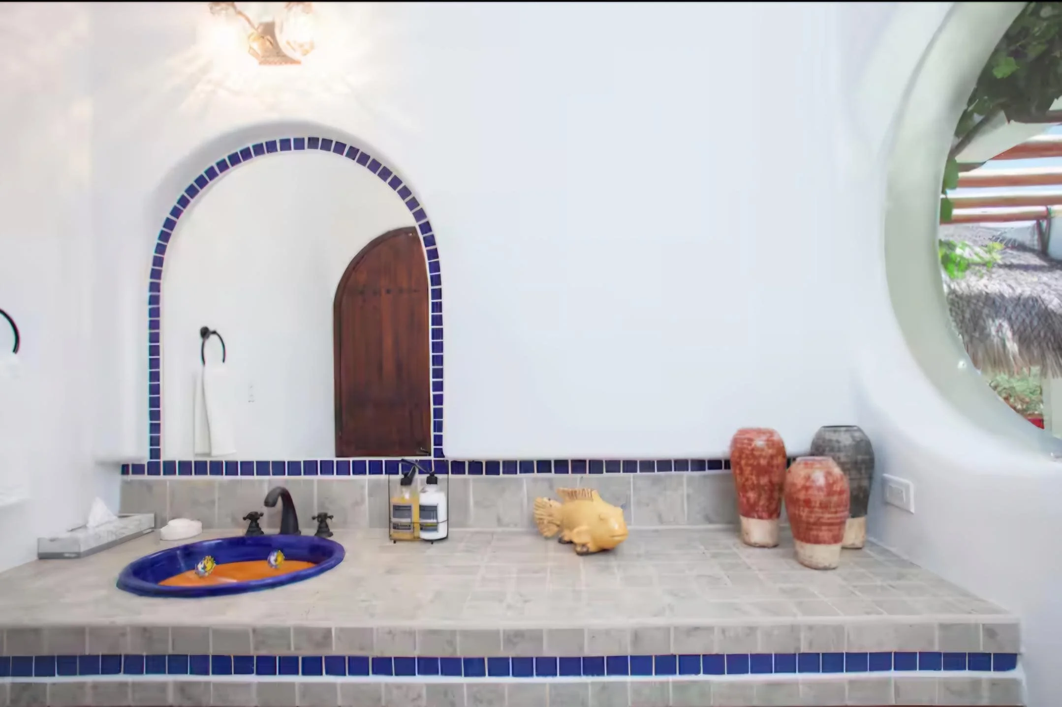 Bathroom with a white wall, arched mirror with blue tile border, and round wooden door. The countertop has a small blue sink with decorative yellow faucet handles, soap dispenser, and toiletries. Two wall-mounted black faucets are beside the mirror. 