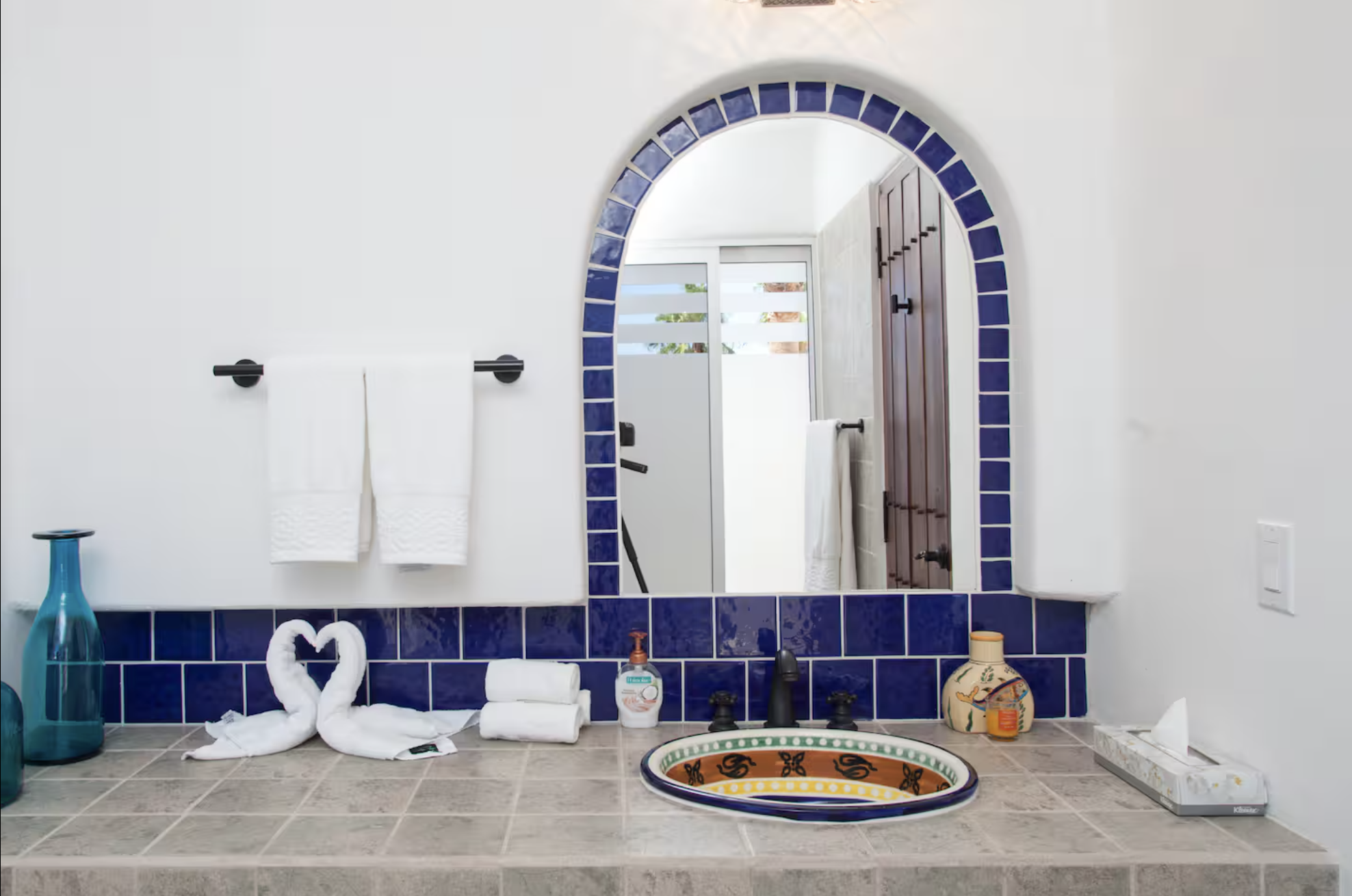 Bathroom countertop with colorful decorative sink, soap dispenser, rolled towels, and decorative folded towels in front of a large mirror with blue tile border, reflecting a door and window with outdoor view.