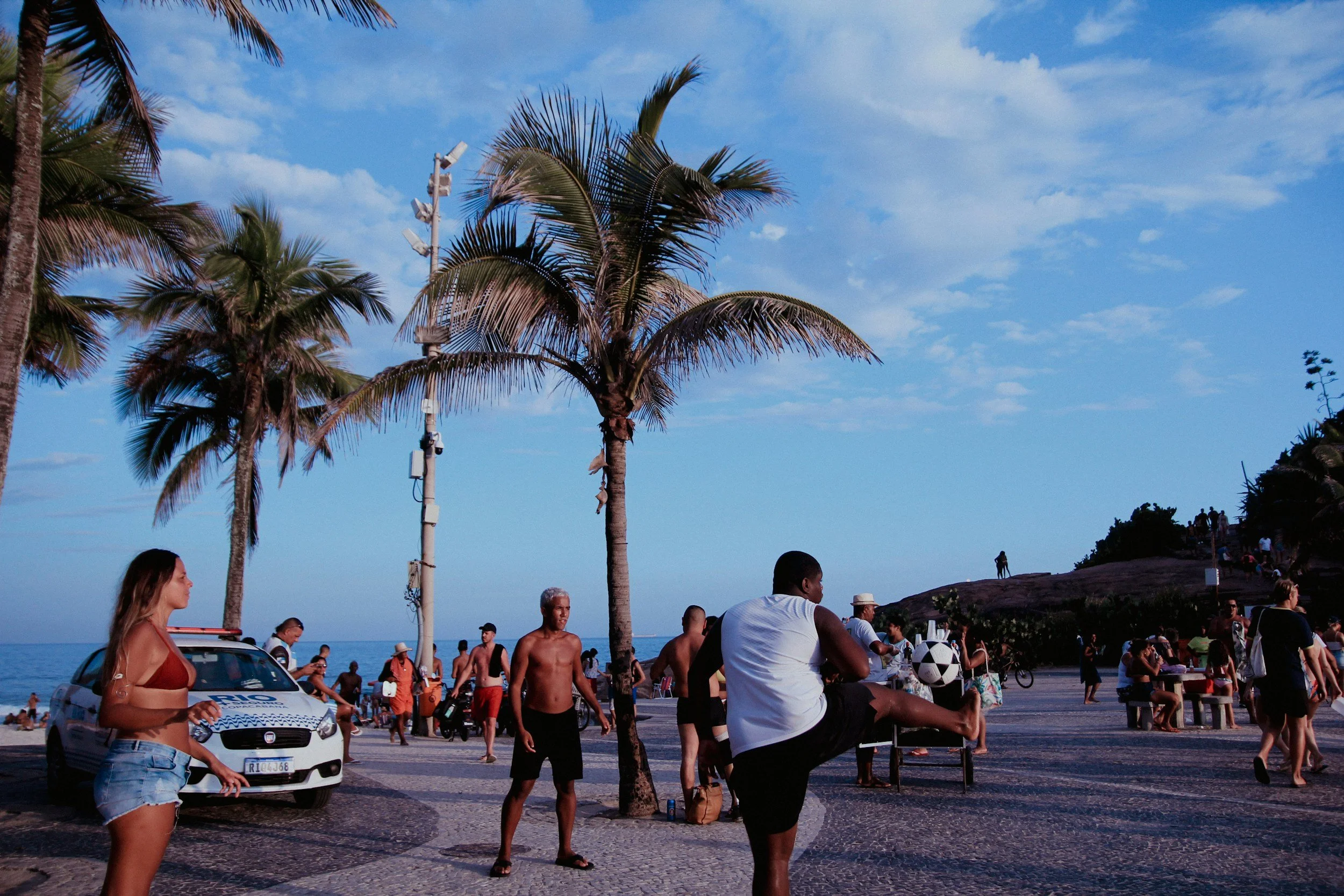 People gathered near palm trees on a beach promenade, with some dancing, walking, and sitting, in a coastal area under a blue sky with scattered clouds.