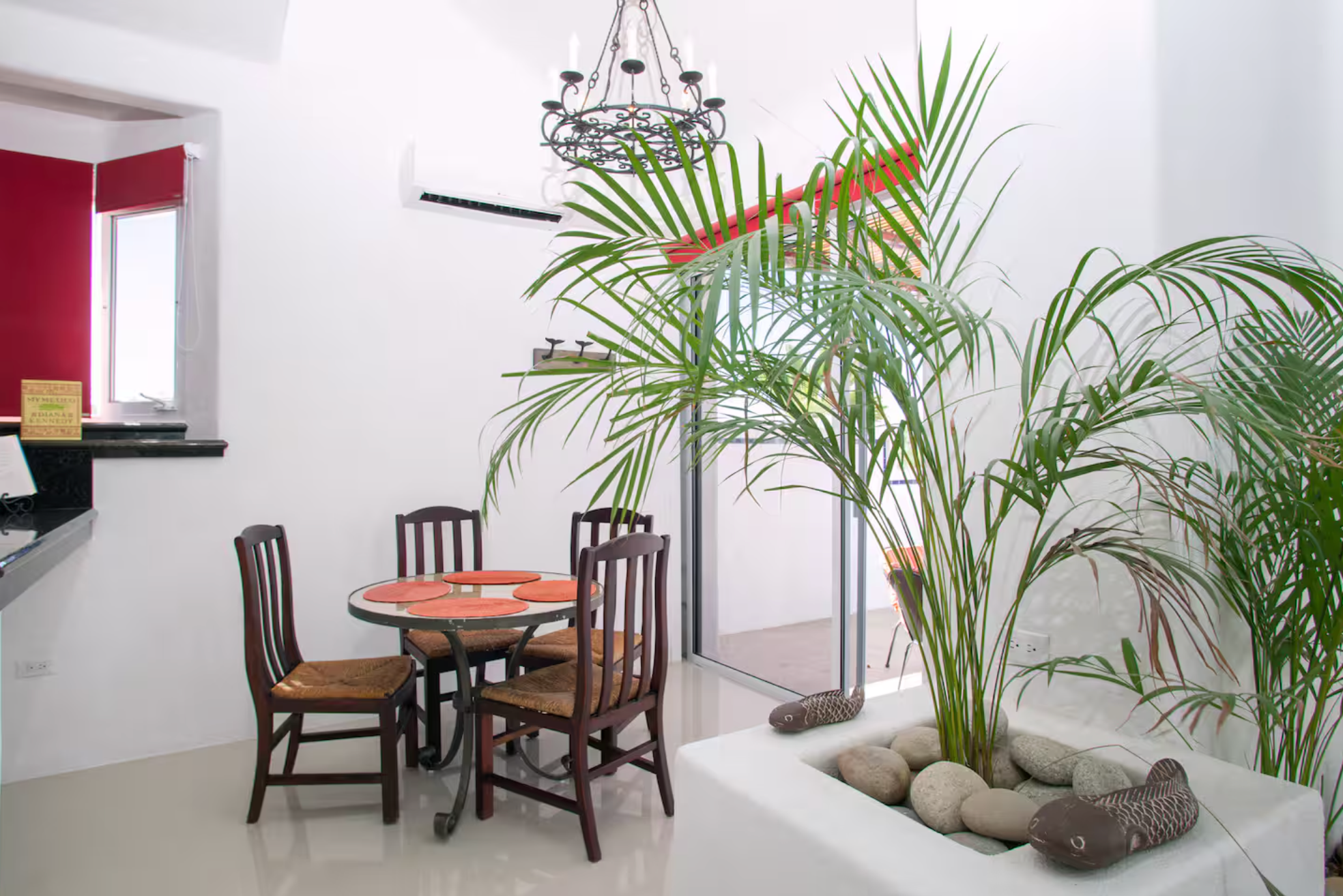 Indoor dining area with a small round table, four wooden chairs with woven seats, large potted palm plant with decorative stone fish ornaments, chandelier hanging from ceiling, and a sliding glass door leading outside.