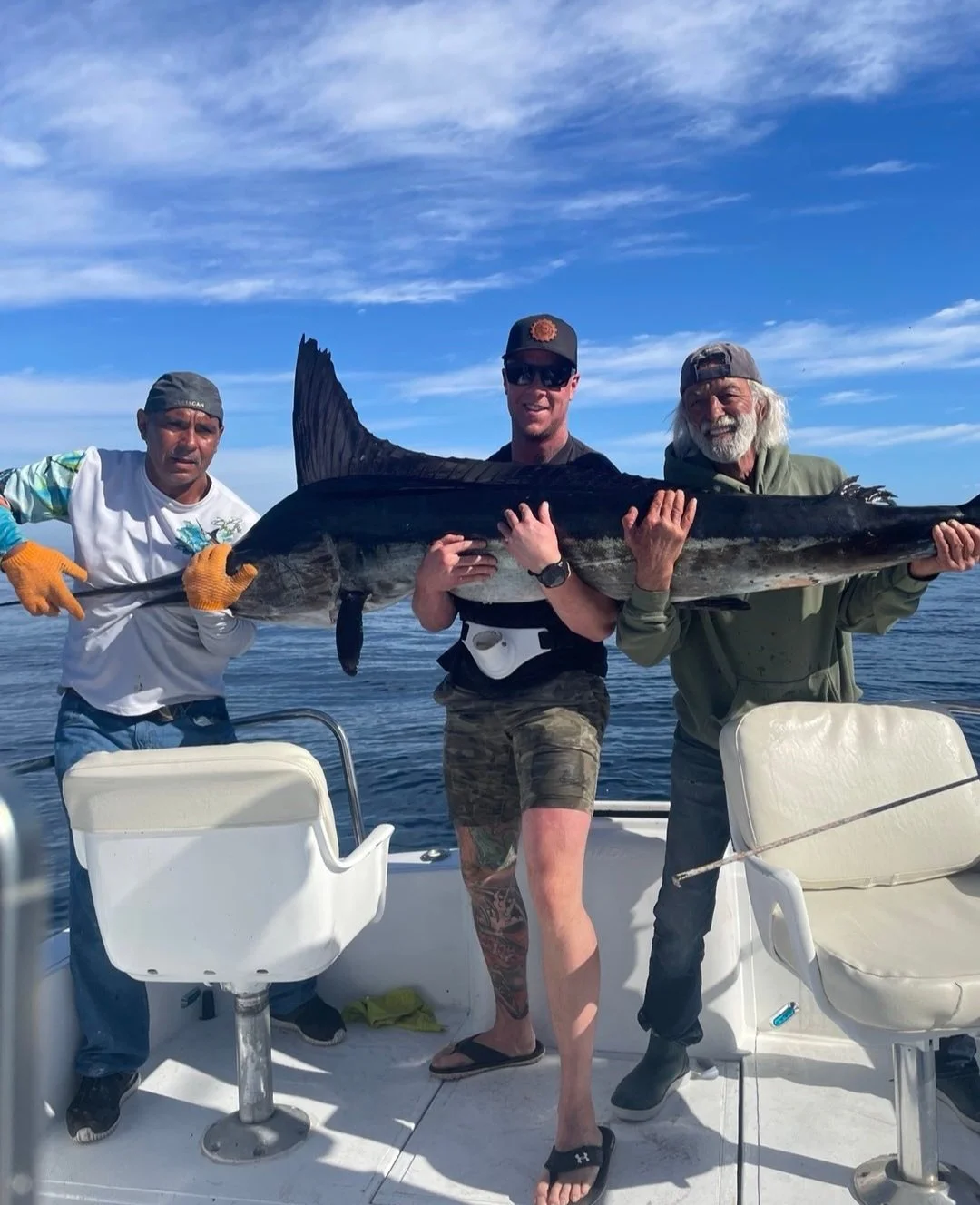 Three men on a boat holding a large fish, likely a halibut, with a blue sky and ocean in the background.