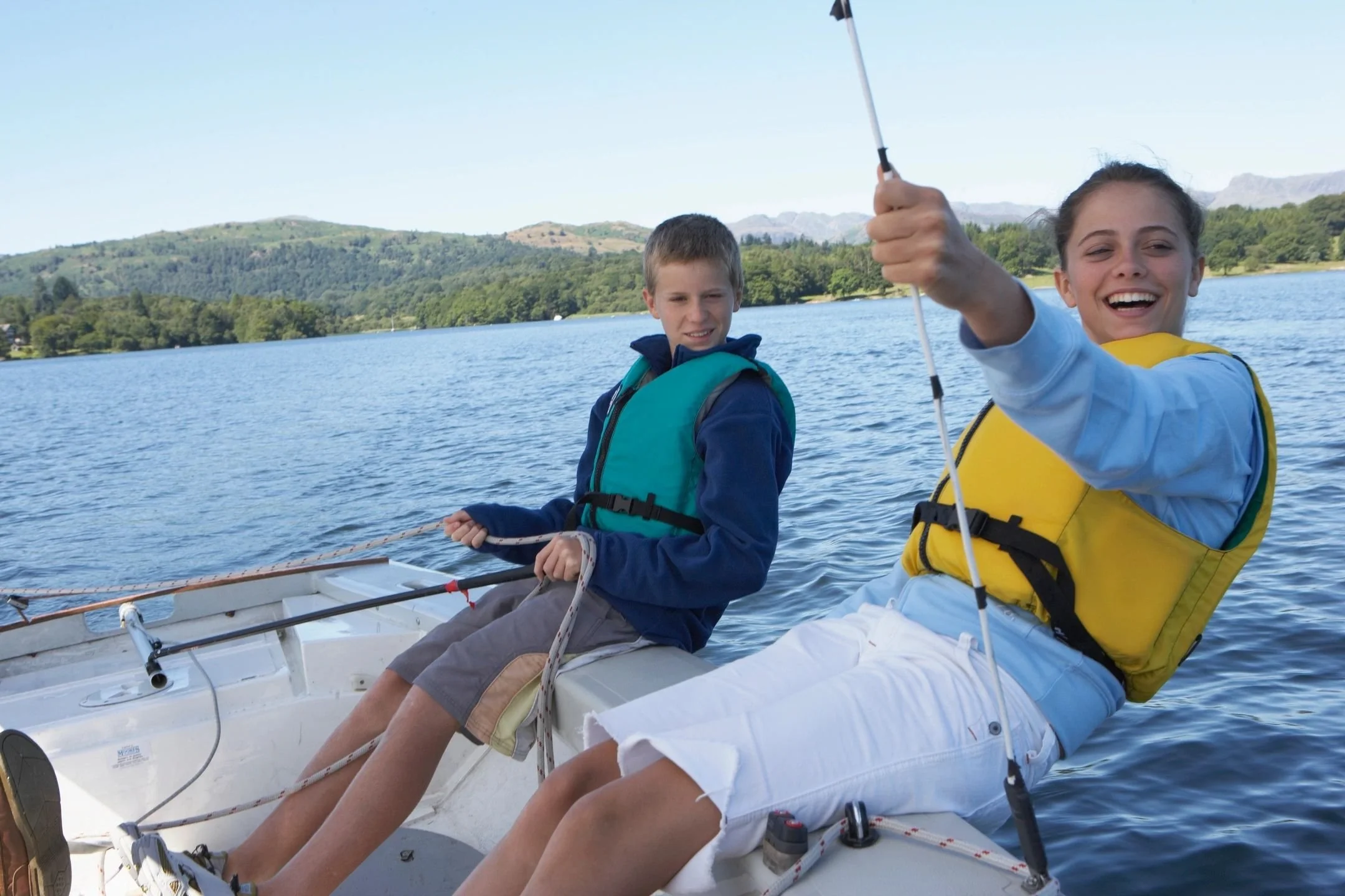 Two children sailing on a lake, smiling and wearing life jackets, with green hills and mountains in the background. The Marlin Hunter Family Boat Charter 
