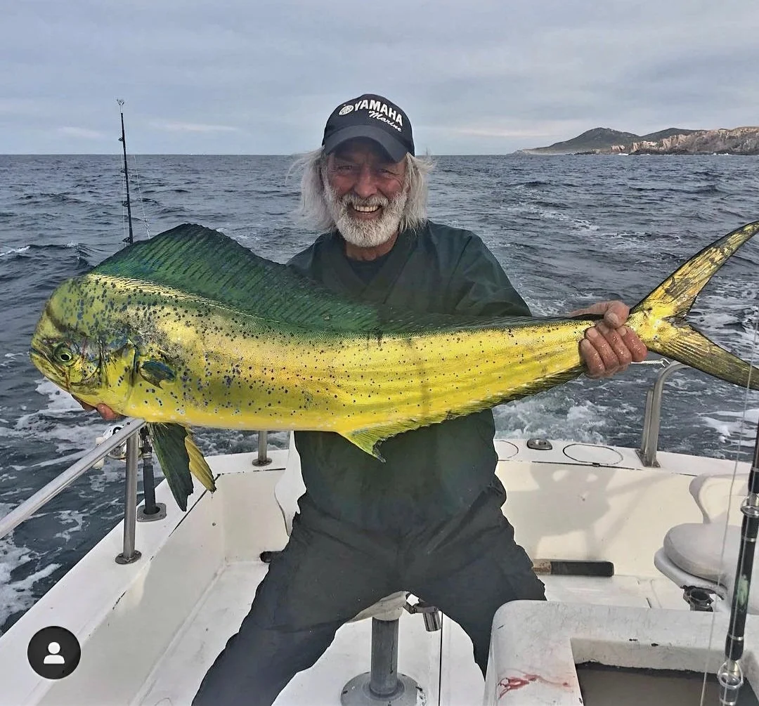 A man with white hair and beard holding a large yellow and green fish on a boat at sea, with a coastline in the background. The Marlin Hunter fishing charters