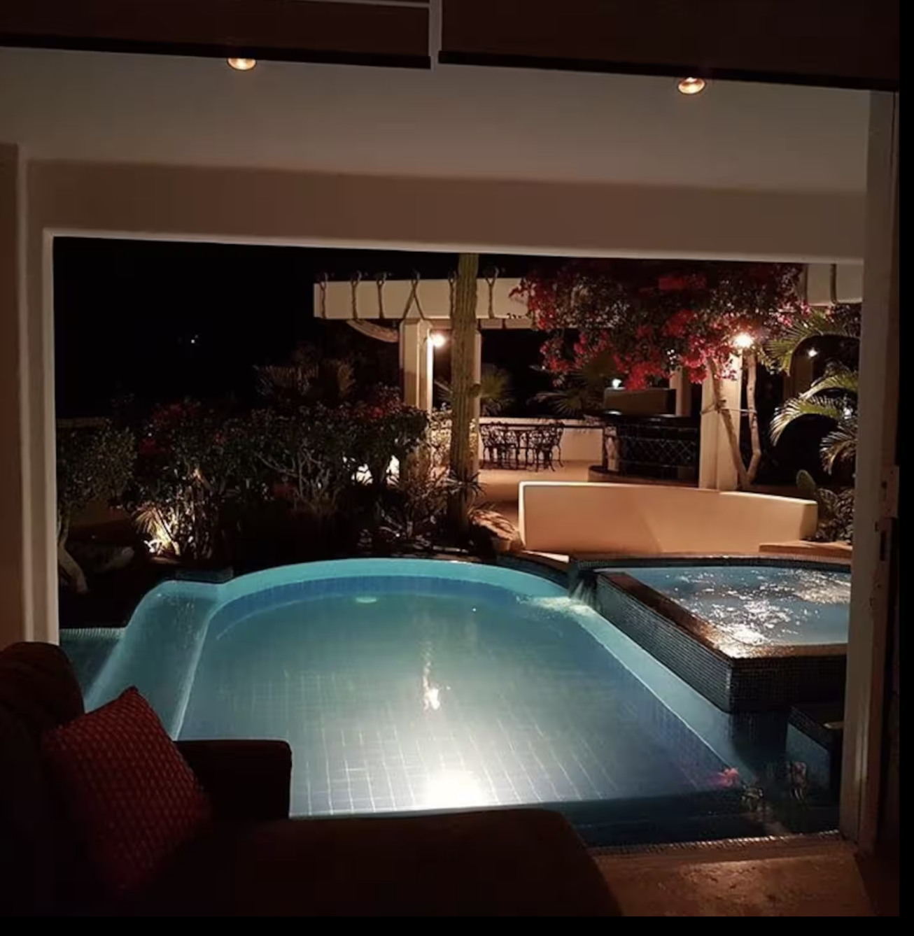 Night view of a luxurious swimming pool with illuminated water, surrounded by lush plants and outdoor seating, under warm lighting.