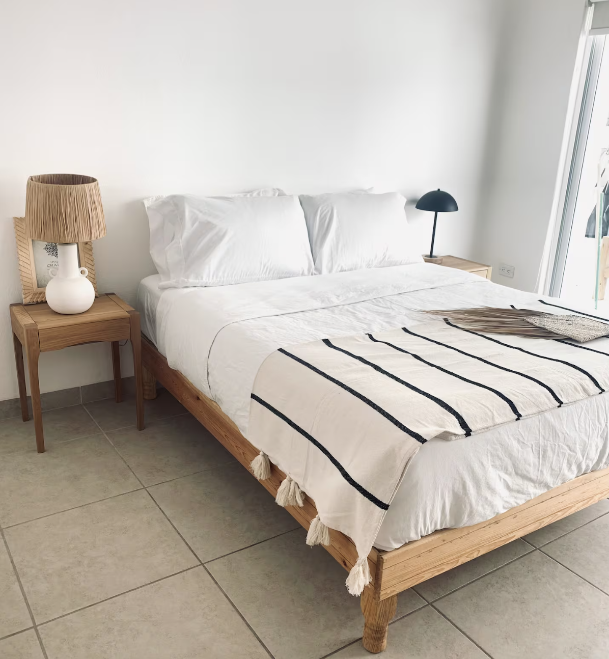 A neatly made bed with white sheets and pillows, a striped beige and black throw blanket at the foot, in a modern bedroom with two wooden side tables, one with a lamp and framed art, and a partially open sliding glass door revealing a balcony or outd