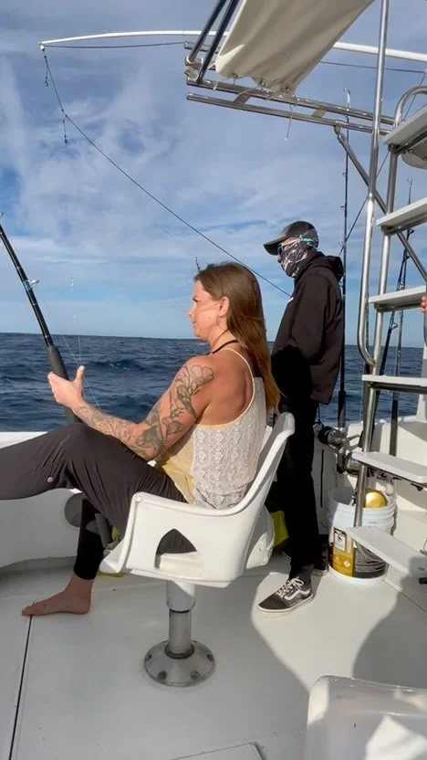 A woman with tattoos sitting on a chair fishing from a boat, with a man standing nearby, both on the open ocean. The Marlin Hunter Fishing Charter Trips