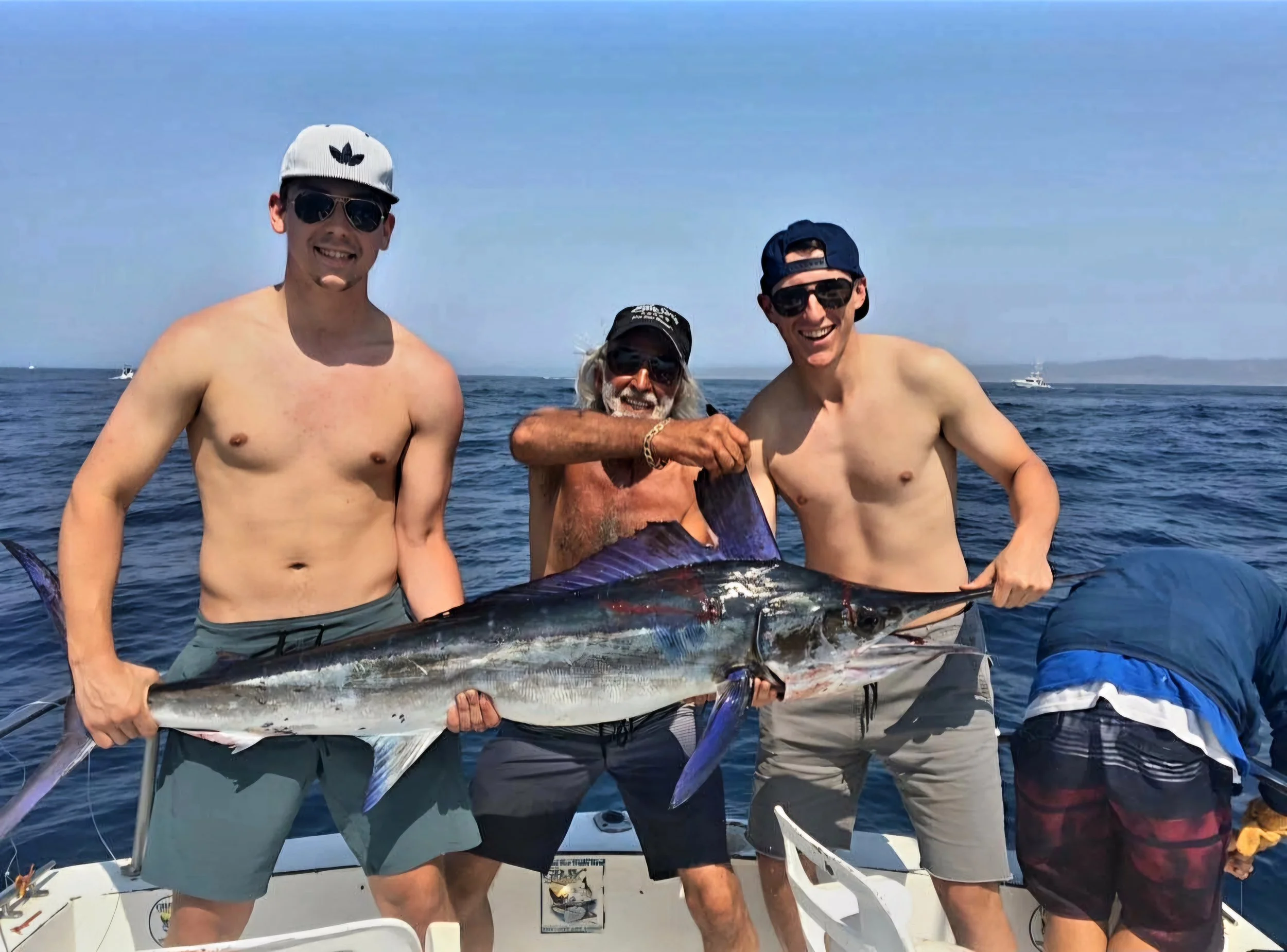 Three shirtless men on a boat holding a large fish, with a man in the middle smiling and wearing sunglasses and a cap, and two other men on either side also smiling, all around on the ocean with boats visible in the background. The Marlin Hunter