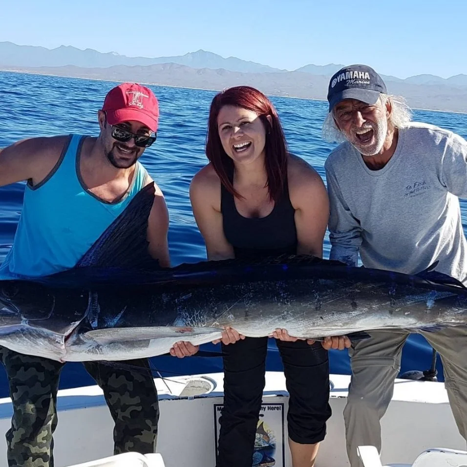 Three people on a boat holding a large fish, with mountains and water in the background. The Marlin Hunter Fishing Charter Day Trip catch of the day