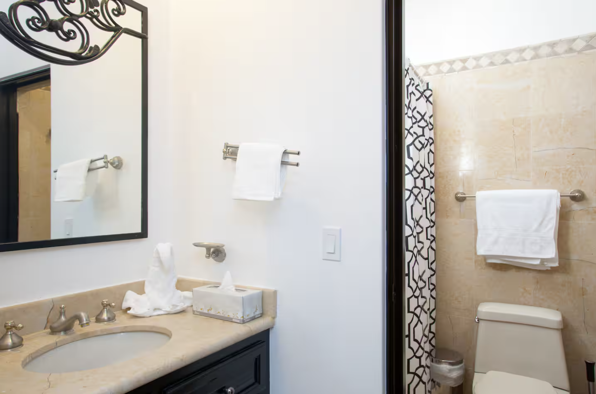 Bathroom with a mirror above a beige marble counter with a sink, soap dispenser, tissue box, and tissue paper. White towels on a wall-mounted bar and a towel ring. A toilet and a shower with a patterned curtain are visible in a separate section.