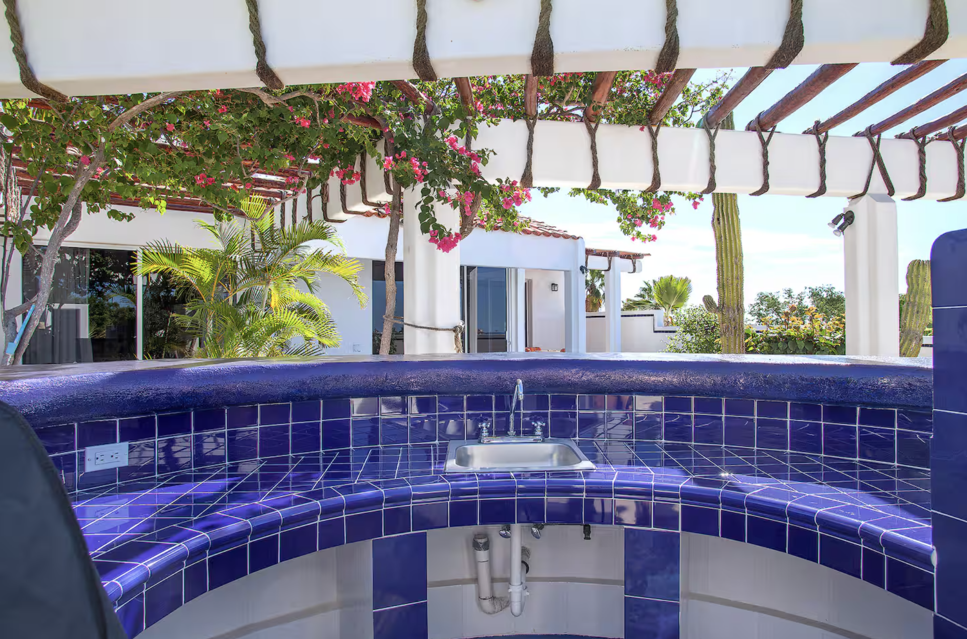 Outdoor hot tub with blue tiles, surrounded by desert plants, trees, and a white patio area under a pergola with pink flowering vines, in a sunny area with clear skies.