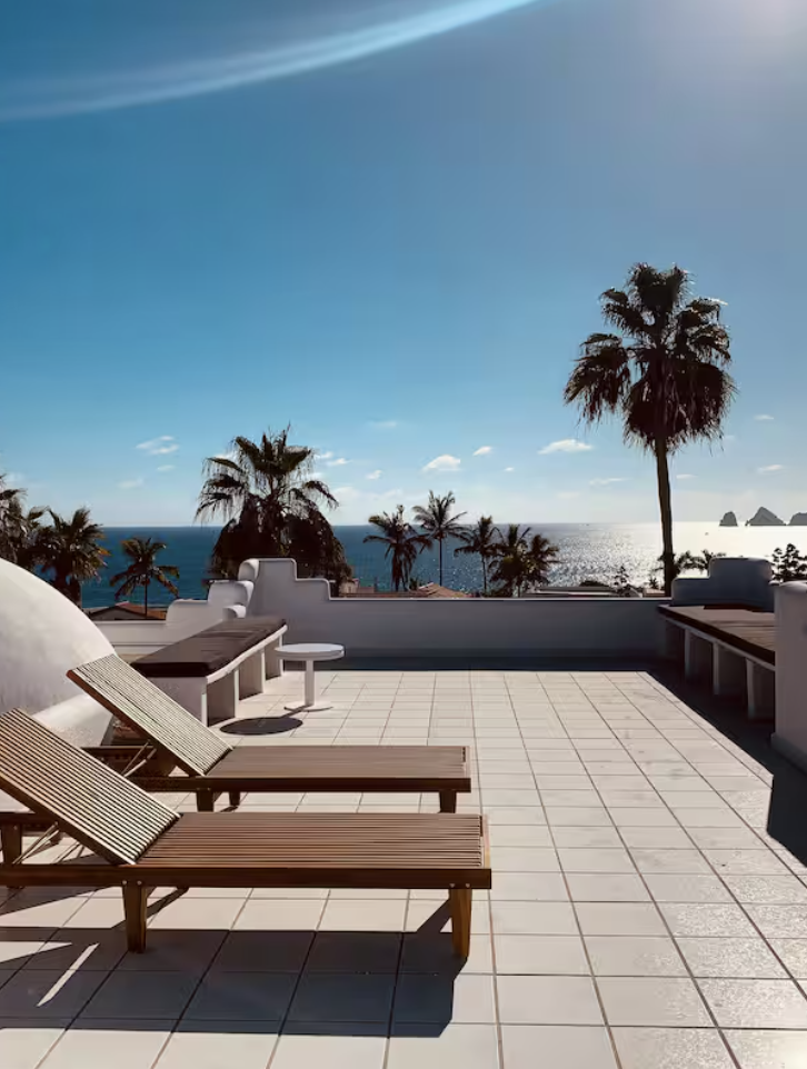 A sunny rooftop terrace with two wooden lounge chairs, a small white side table, and a view of palm trees and the ocean under a blue sky.