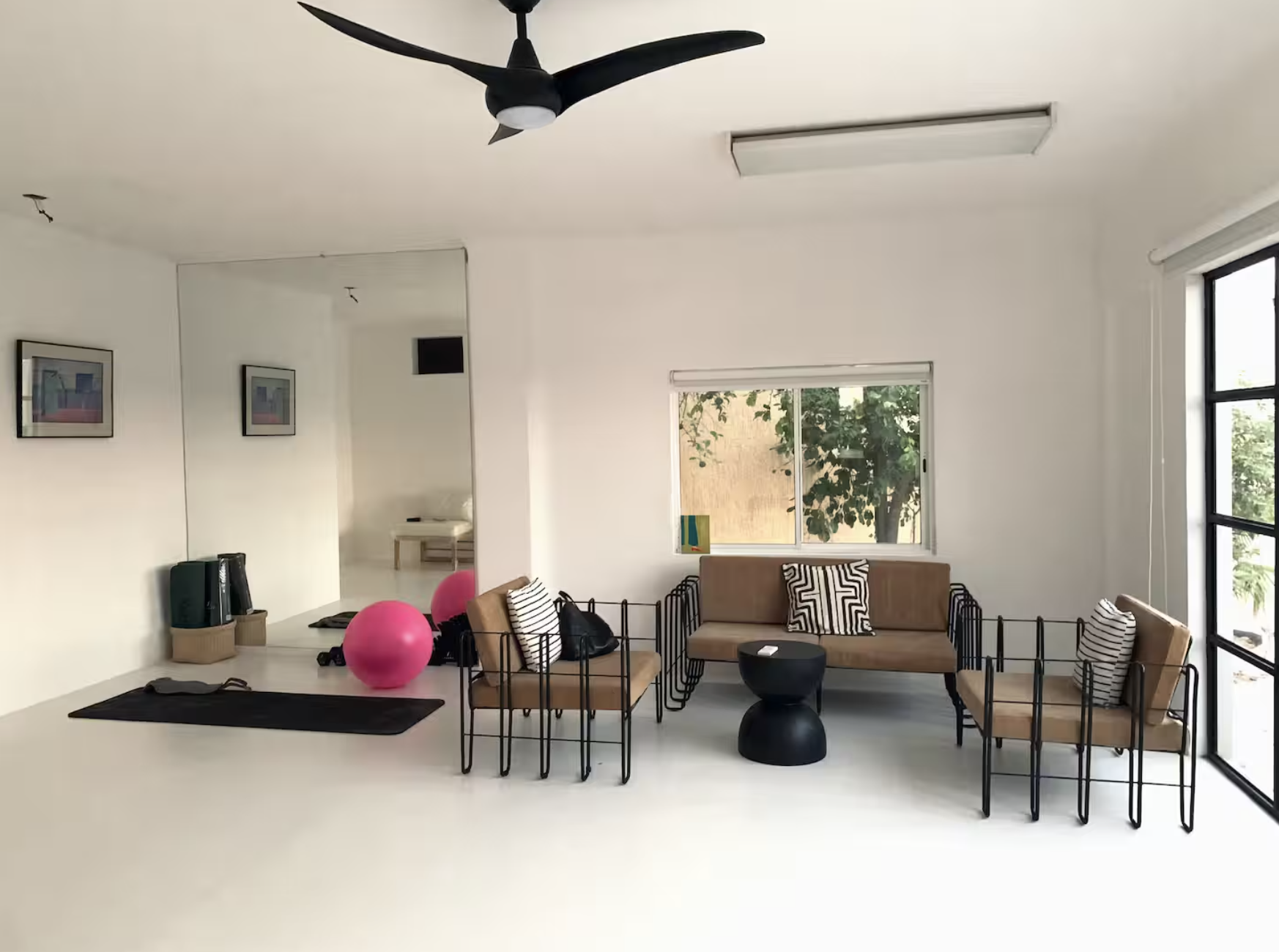 Living room with white walls, a black ceiling fan, a large window, beige seating with black metal frames, patterned throw pillows, a black round side table, and exercise balls and mats on the floor.