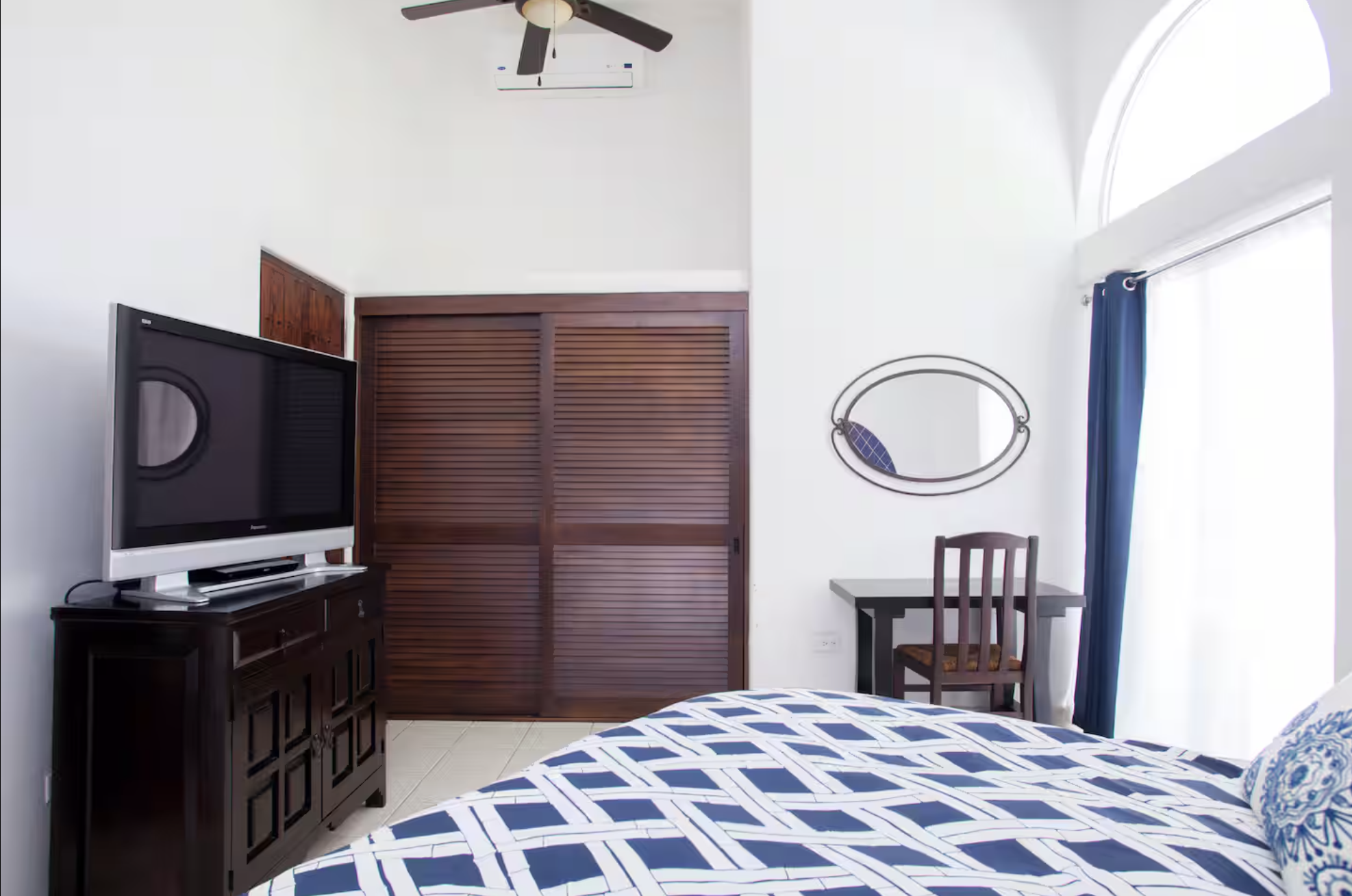 A bedroom with a dark wood dresser holding a flat-screen TV, a bed with a blue and white patterned quilt, a small black desk with a wooden chair with a striped cushioned seat, a decorative mirror on the wall, a sliding glass door with blue curtains, 