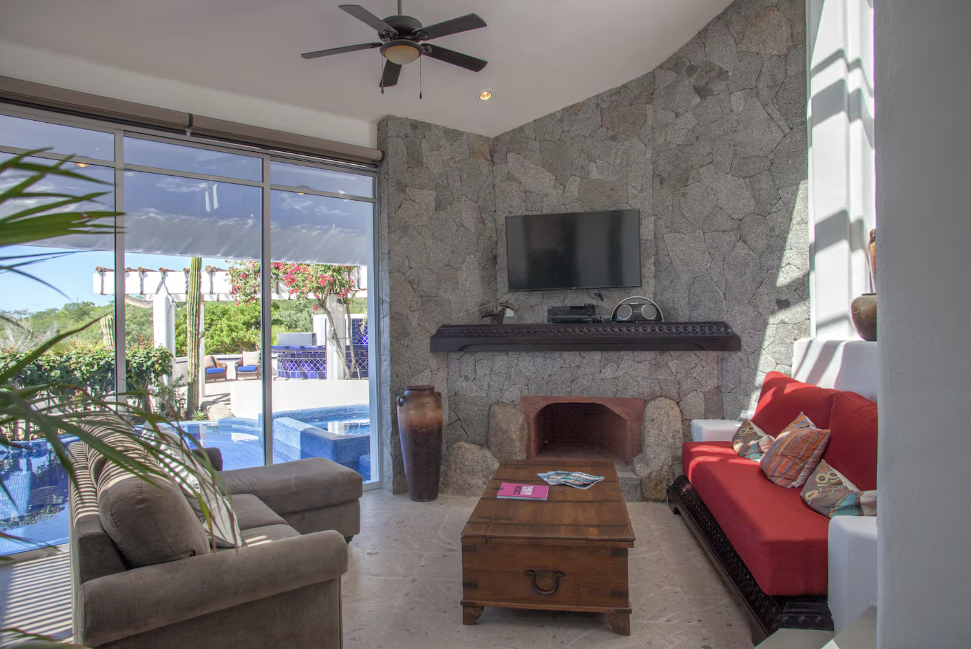 Living room with stone accent wall, television, fireplace, red and beige sofas, wooden coffee table, indoor plants, large glass sliding doors opening to an outdoor pool area with lounge chairs and cacti.