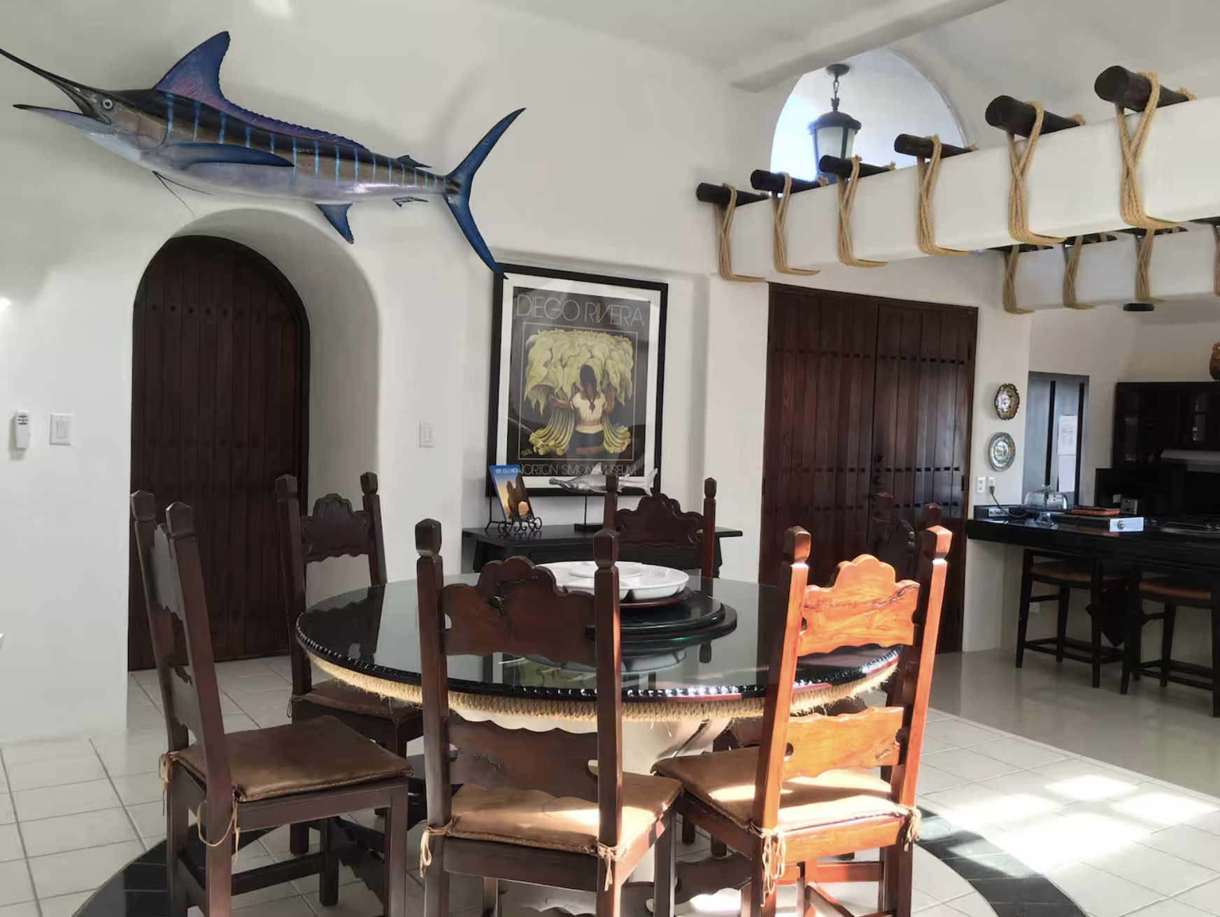 Dining room with a round black table surrounded by six wooden chairs with beige cushions, a large marlin mounted on the wall, a framed poster of Diego Rivera, and wooden Double doors.
