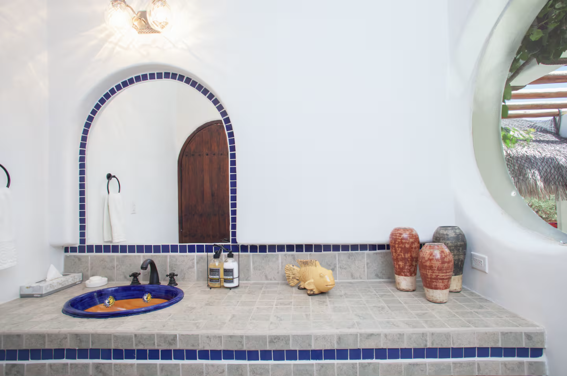 Bathroom vanity with ceramic fish-shaped sink, decorative vases, soap dispensers, and a mirror framed with blue tiles, situated in a room with a curved wall.