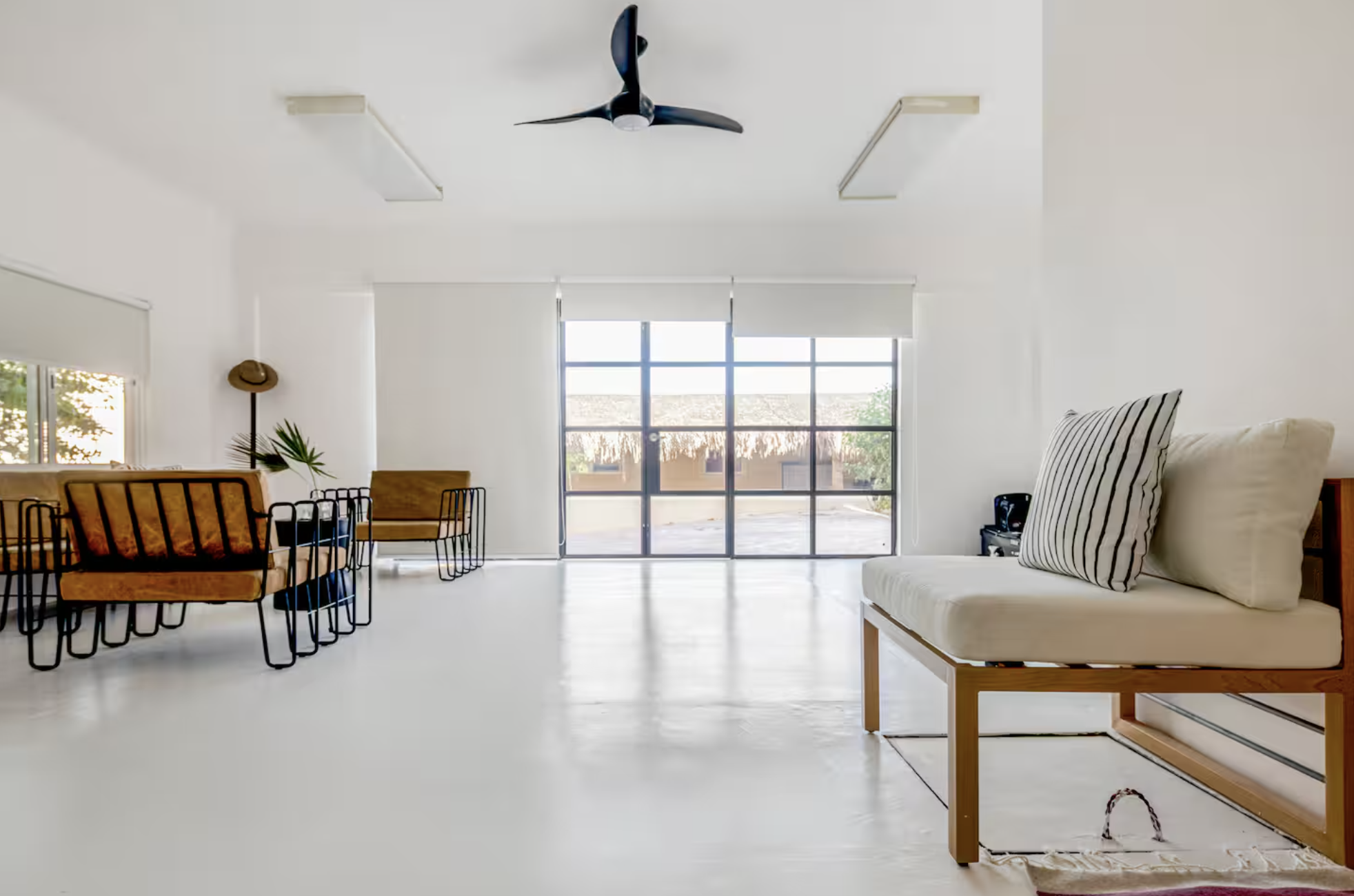 Minimalist living room with white walls and flooring, large glass door with blinds, black ceiling fan, wooden chairs with orange cushions, a small side table, green plant, and a white sofa with striped pillows.