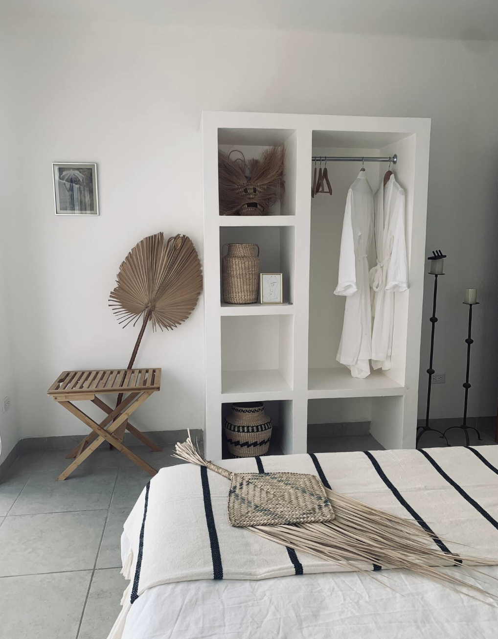 Decorative bedroom corner with a white open wardrobe containing white robes, wicker baskets, and picture frames. A small wooden foldable table with a large dried palm leaf is on the left, a framed picture on the wall, and a bed with a white and black